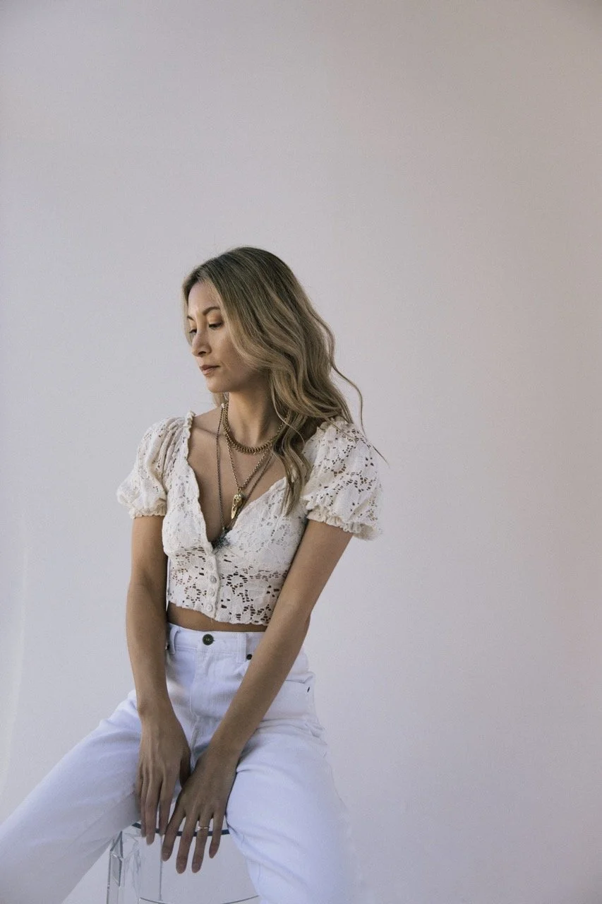 A woman with wavy blonde hair wearing a white lace crop top and white pants, sitting against a plain light-colored background.
