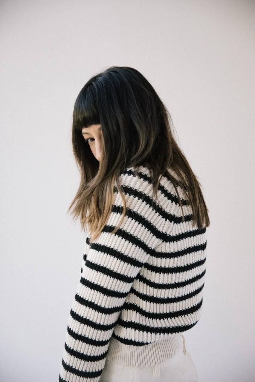 A young woman with long, wavy brown hair with blonde highlights, wearing a black and white striped sweater, standing against a plain light background.