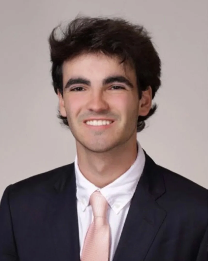 A young man with dark curly hair, wearing a black suit, white shirt, and light pink tie, smiling at the camera against a plain background.