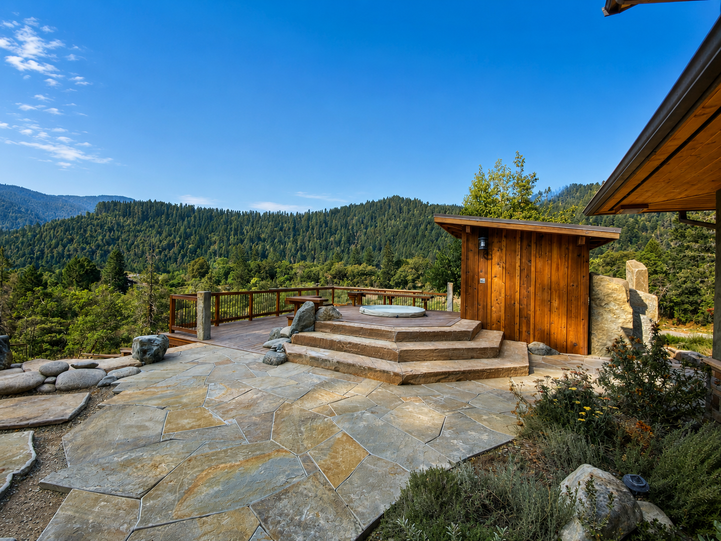Outdoor deck with hot tub, natural stone flooring, wooden privacy wall, overlooking lush green forested mountains under clear blue sky.