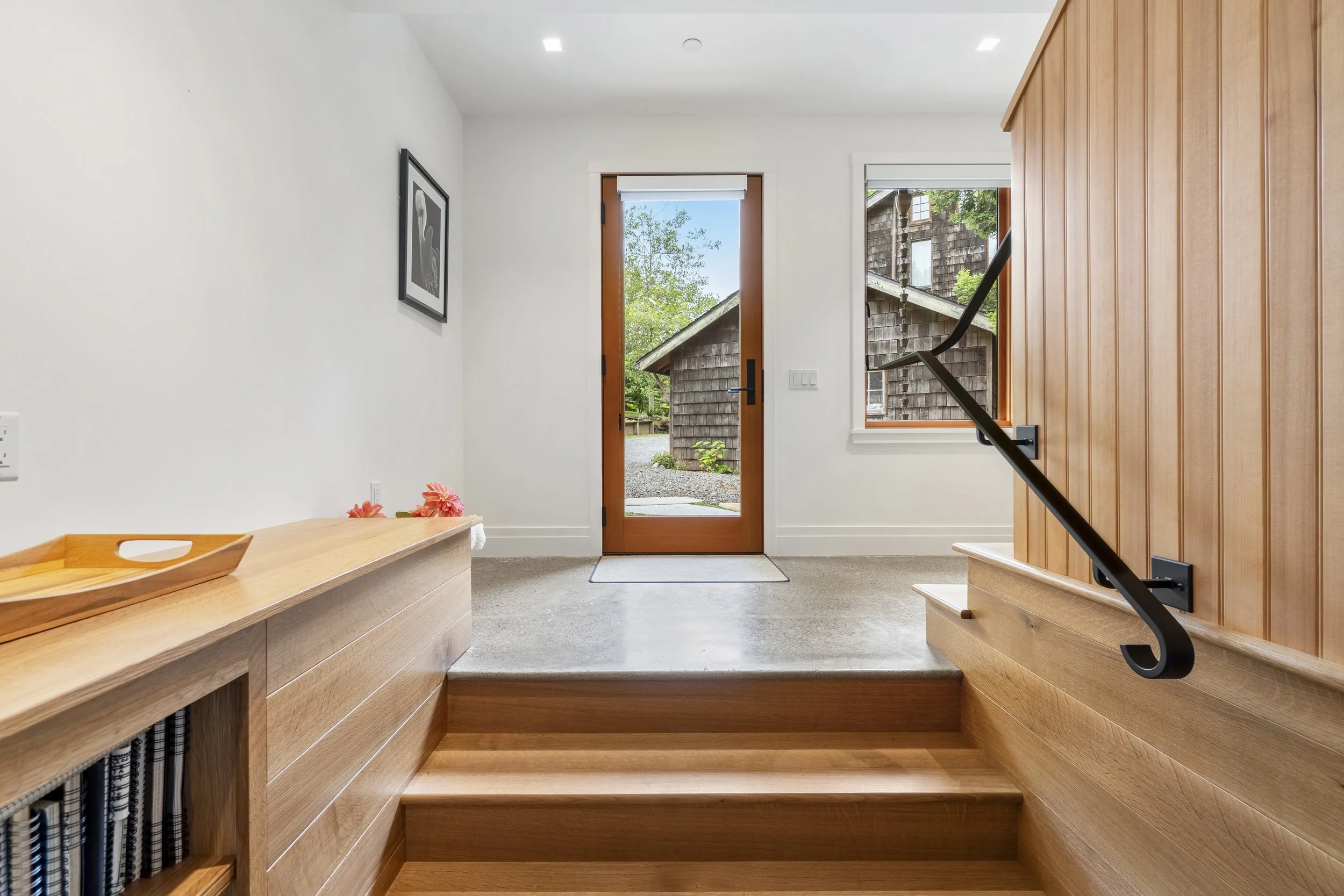 Entryway with wooden flooring and built-in wooden benches on both sides, a glass door, and windows showing a house exterior and trees outside.