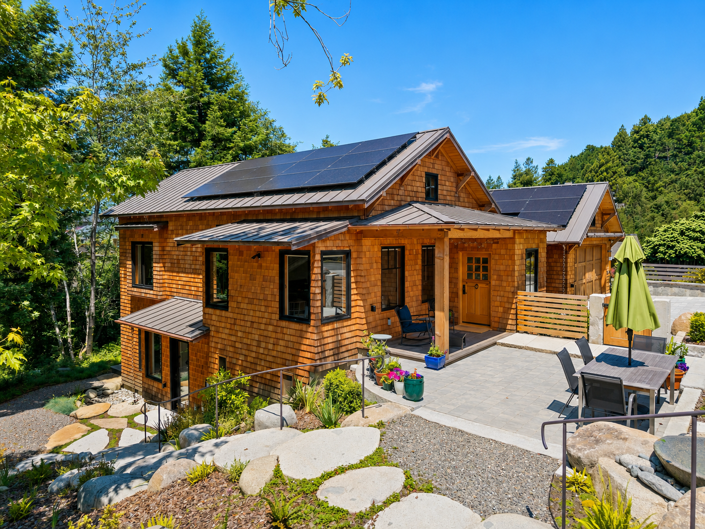 A modern cedar-shake house with a metal roof featuring solar panels, surrounded by lush greenery and a patio with outdoor furniture, umbrellas, and potted plants.