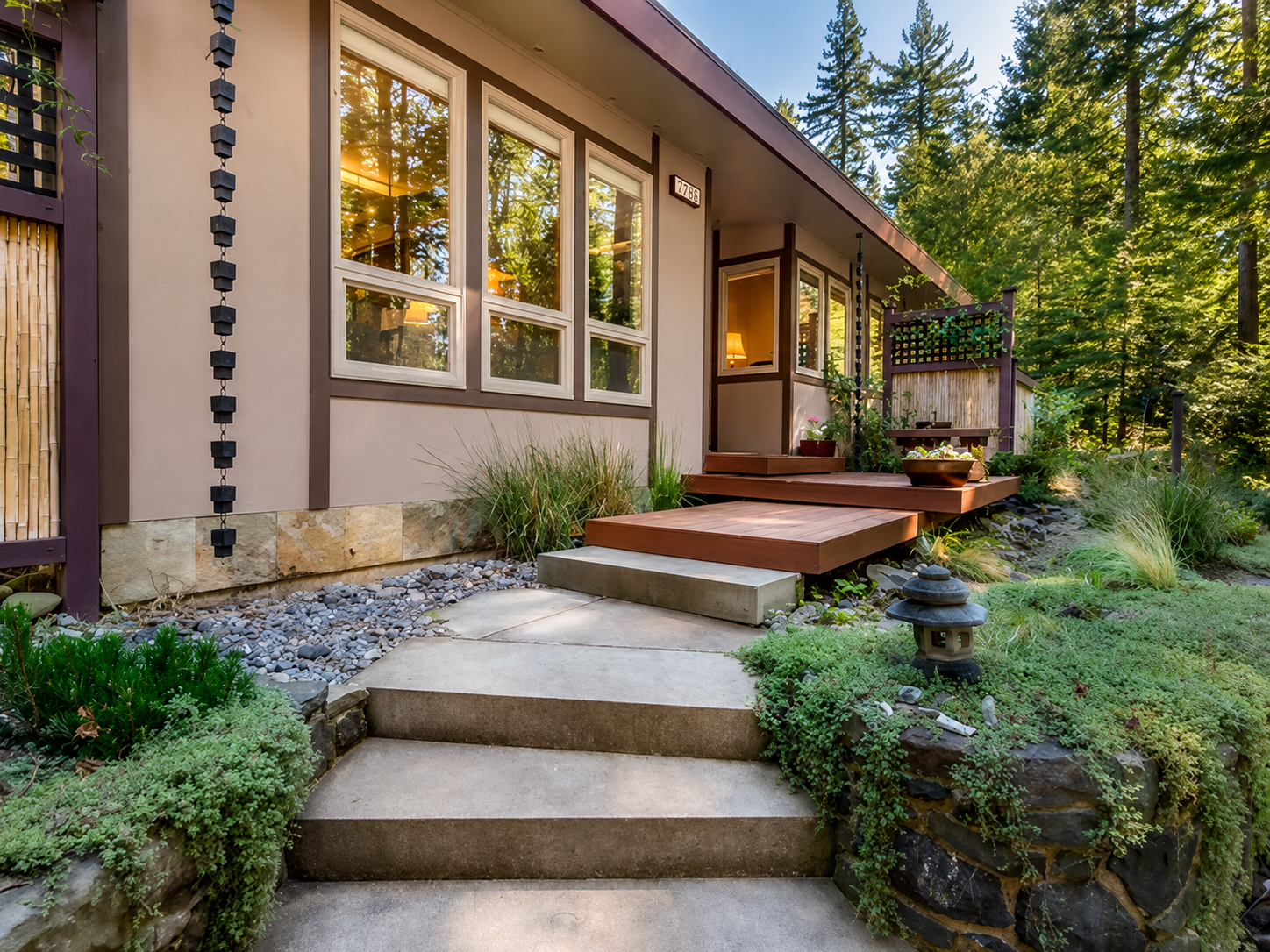 Front porch of a house with concrete steps leading to a wooden deck, surrounded by greenery, with large windows reflecting trees, and a wooded background.