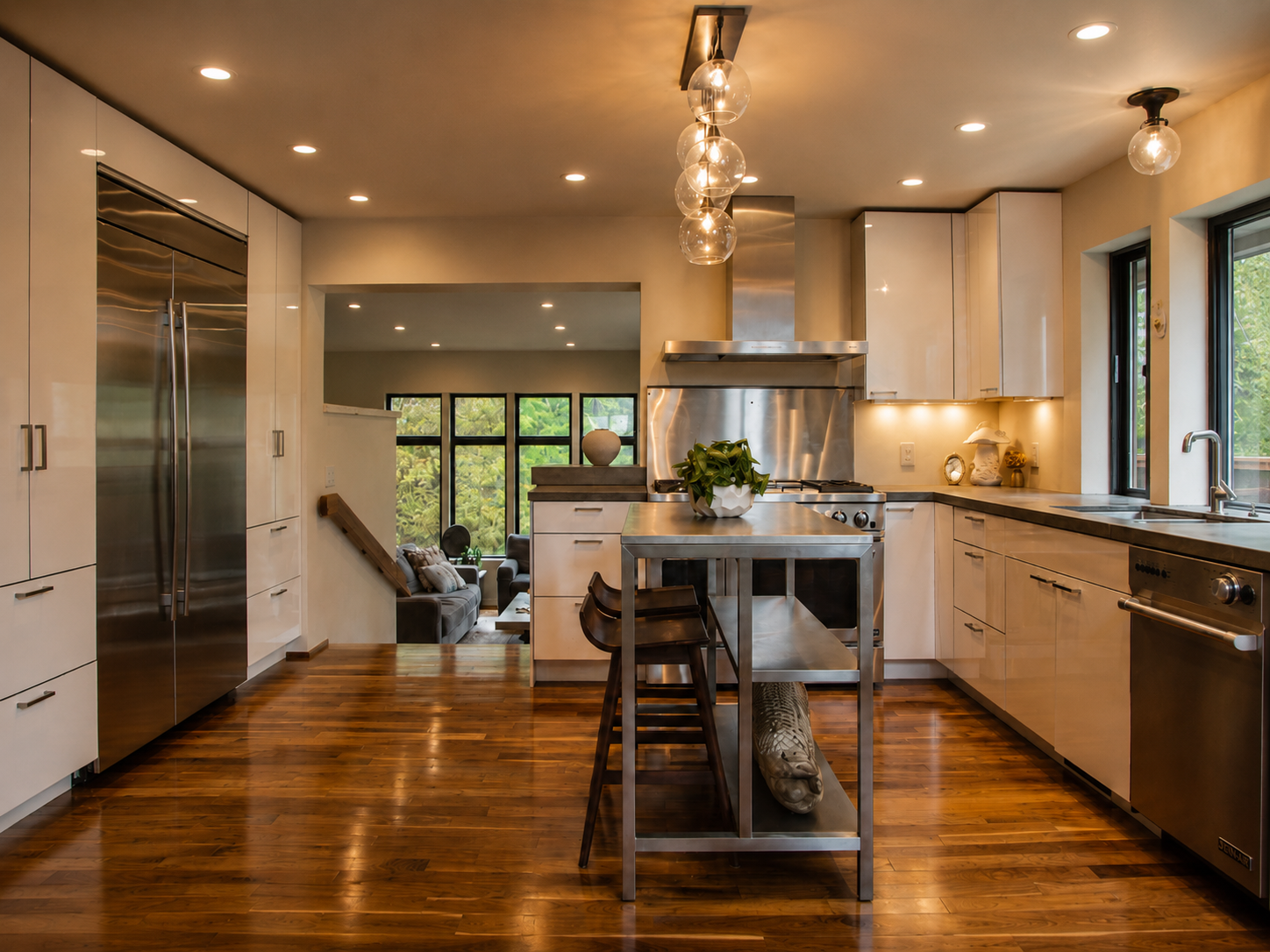 Modern kitchen with white cabinets, stainless steel appliances, wooden flooring, and a view into a living room with large windows and greenery outside.