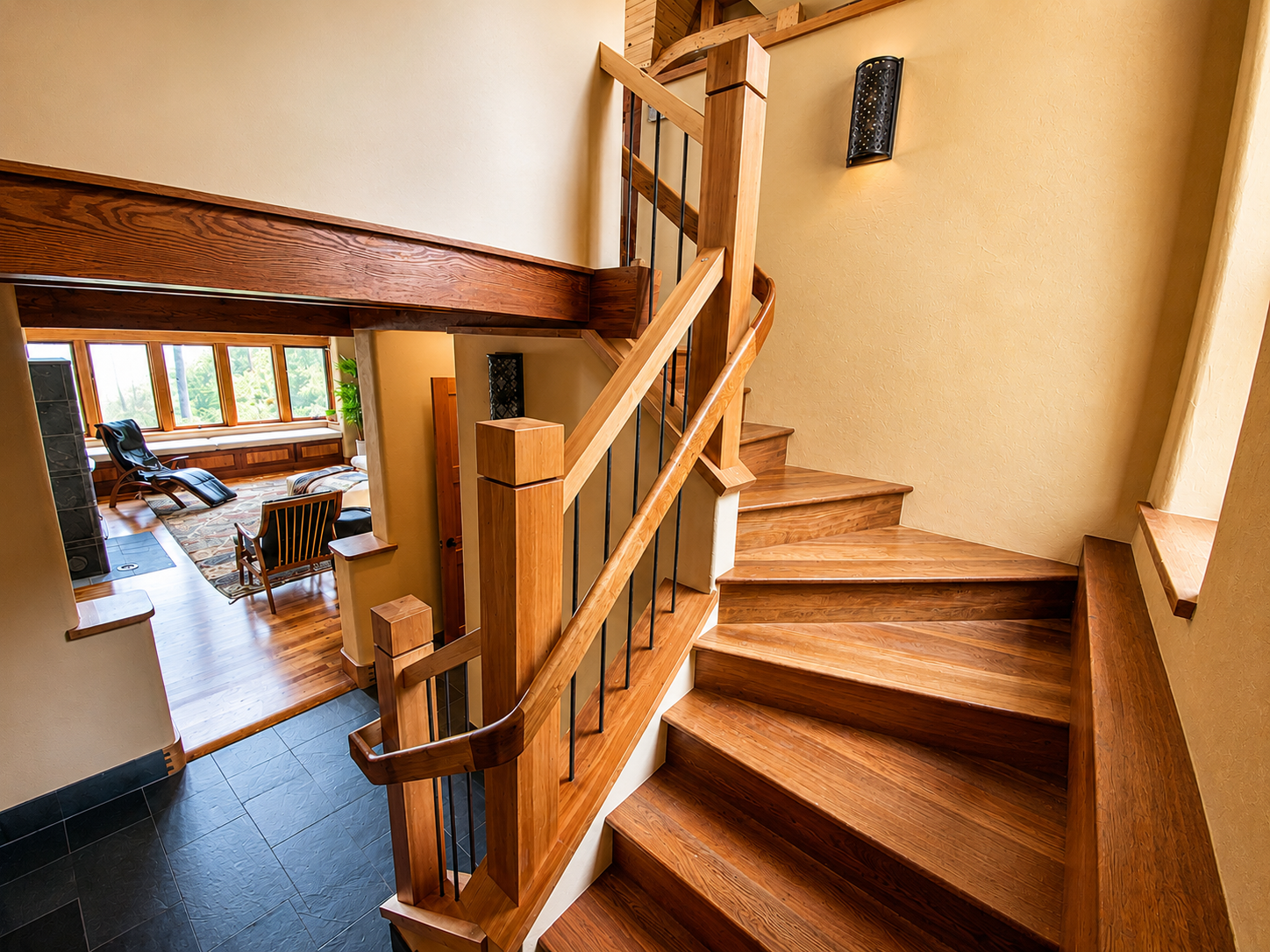 Wooden staircase with black metal railing leading to a second floor in a cozy home interior, with a living room visible below through an open area, featuring large windows and wooden furniture.