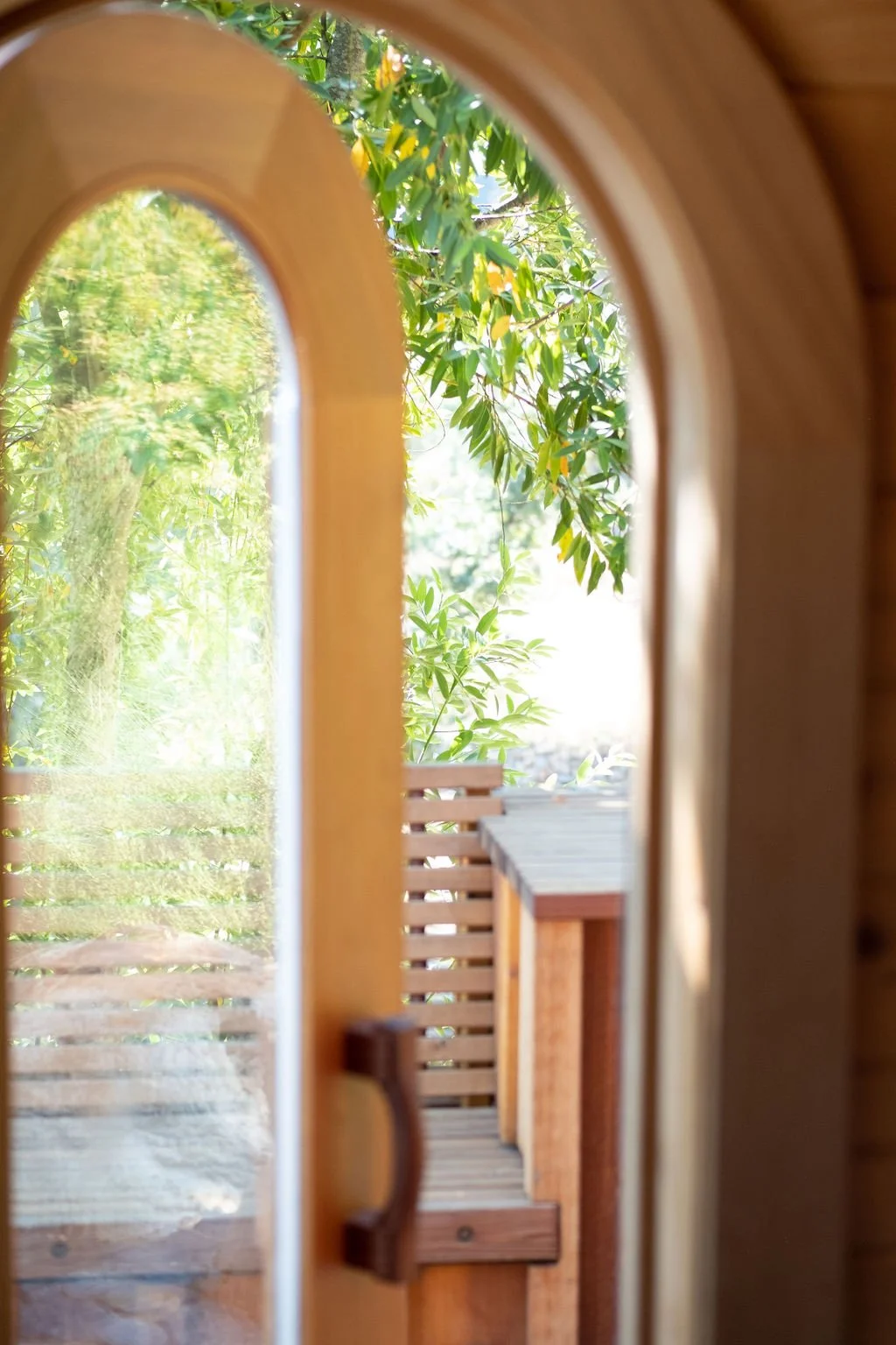 Looking through a small arched window with wooden framing at an outdoor scene consisting of a wooden deck, green trees, and sunlight.