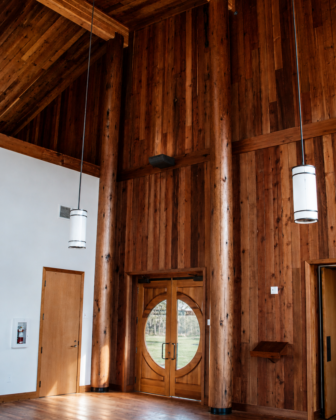 Interior view of a wooden building with high ceilings, hanging cylindrical lights, a rounded wooden door with glass panels, and a small wooden shelf on the wall.