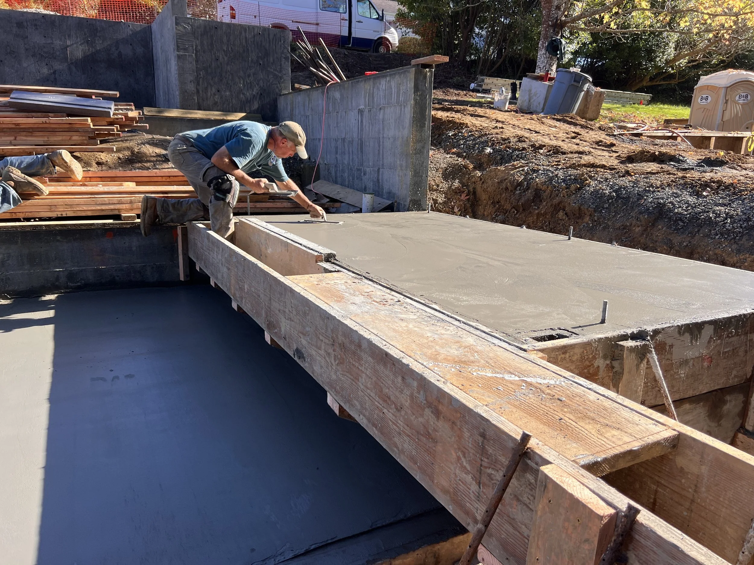 A construction worker applying a fresh layer of concrete on a foundation with a trowel, surrounded by construction materials and equipment at an outdoor site.