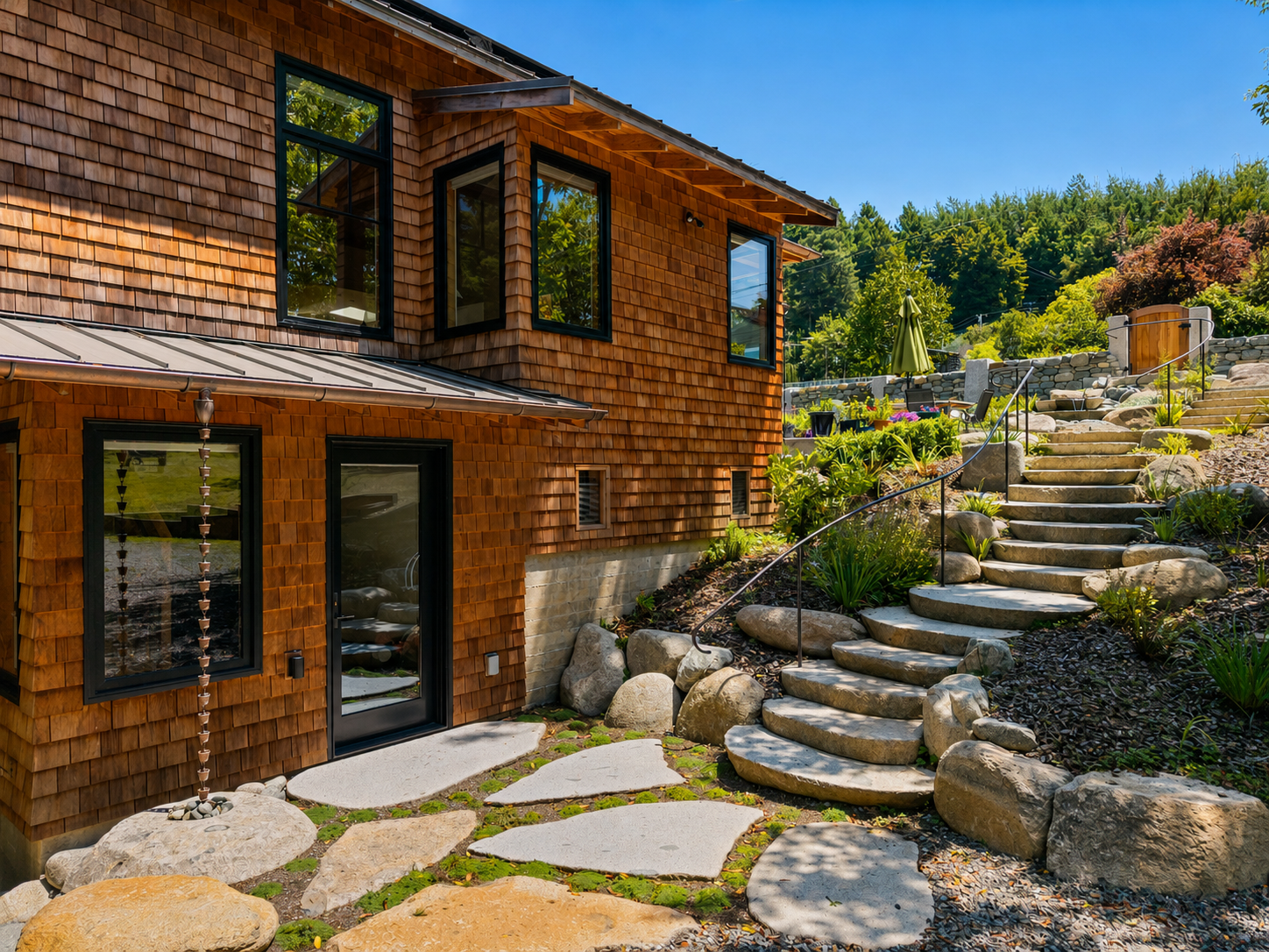 Exterior view of a modern house with wooden shingle siding, multiple windows, and a sliding glass door. Stone steps and a curved pathway lead up a landscaped hillside with plants and rocks, set against a backdrop of trees and a clear blue sky.