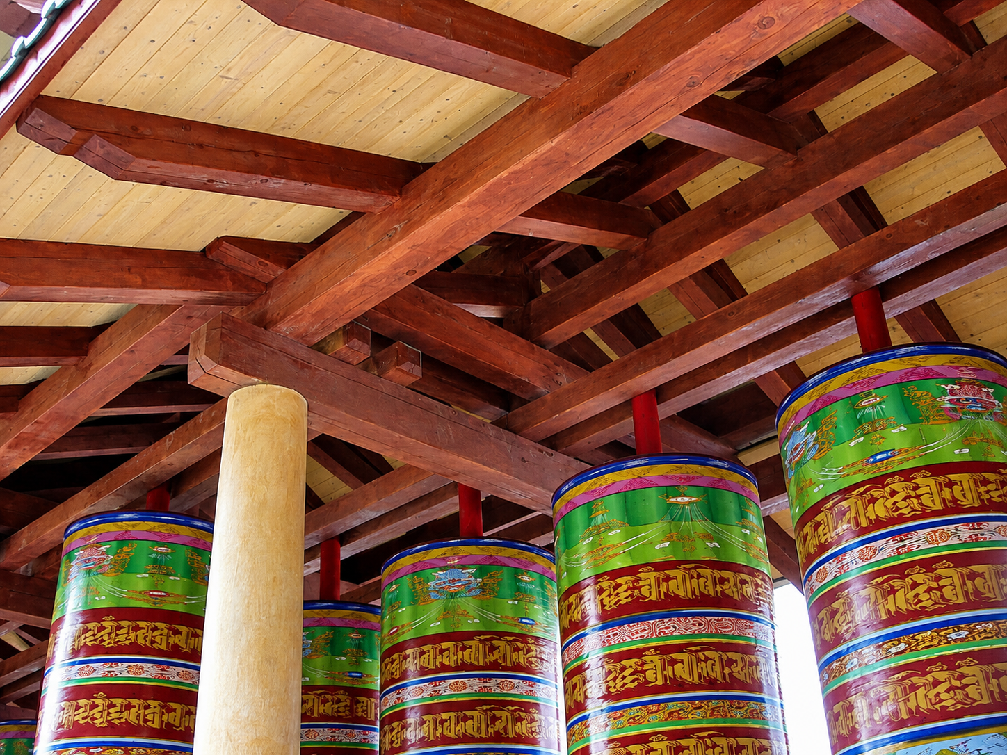 Multiple colorful Tibetan prayer wheels hanging from a wooden temple ceiling with intricate wood beams and a light-colored pillar in the foreground.