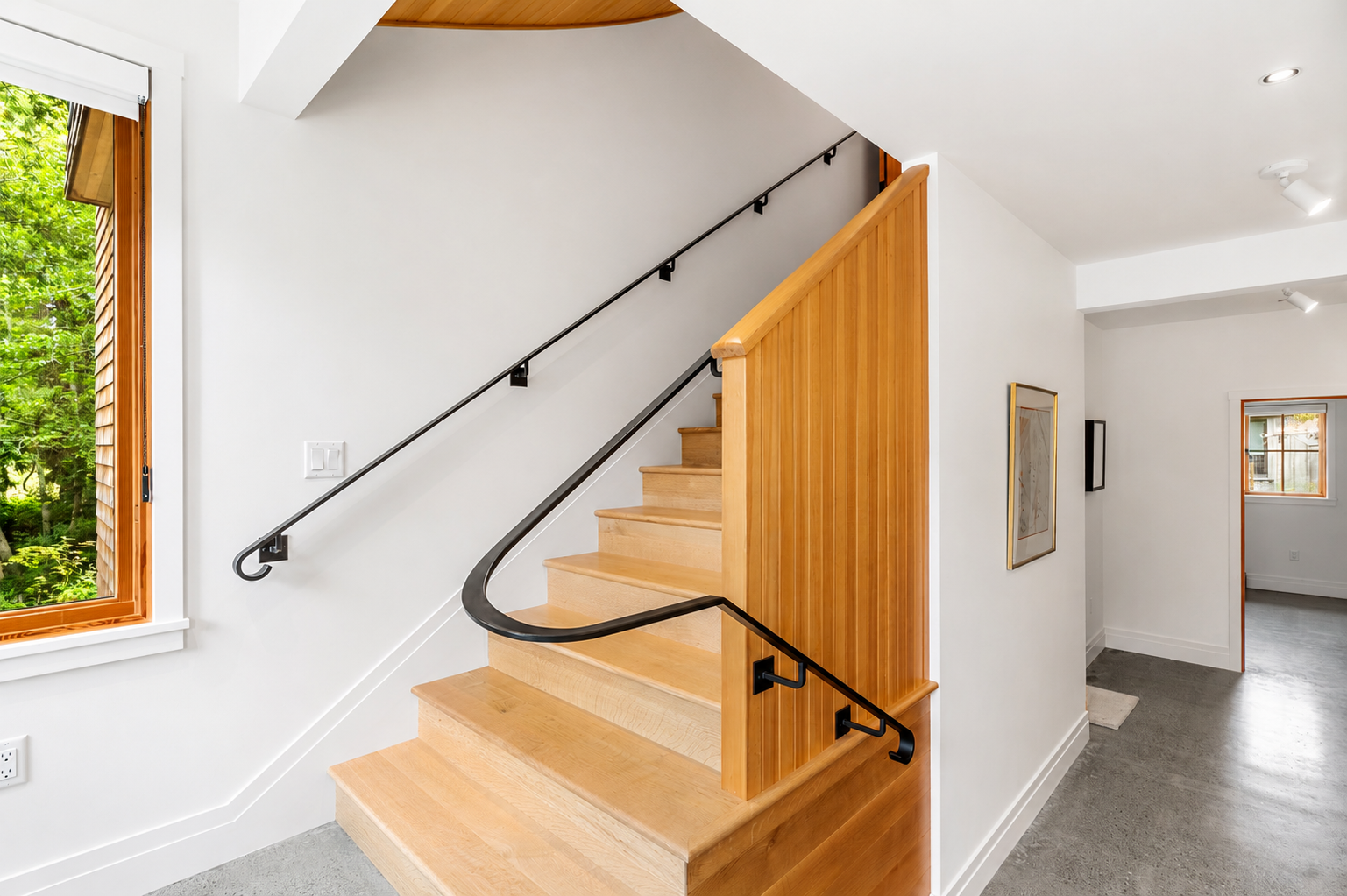 Interior of a modern house showing a wooden staircase with black handrail, white walls, a window with trees outside, and a hallway with framed artwork.