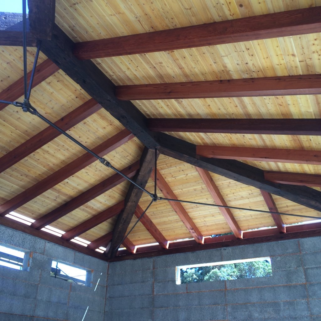 Interior view of a building under construction with a wooden roof and exposed beams, and cinder block walls with small windows.