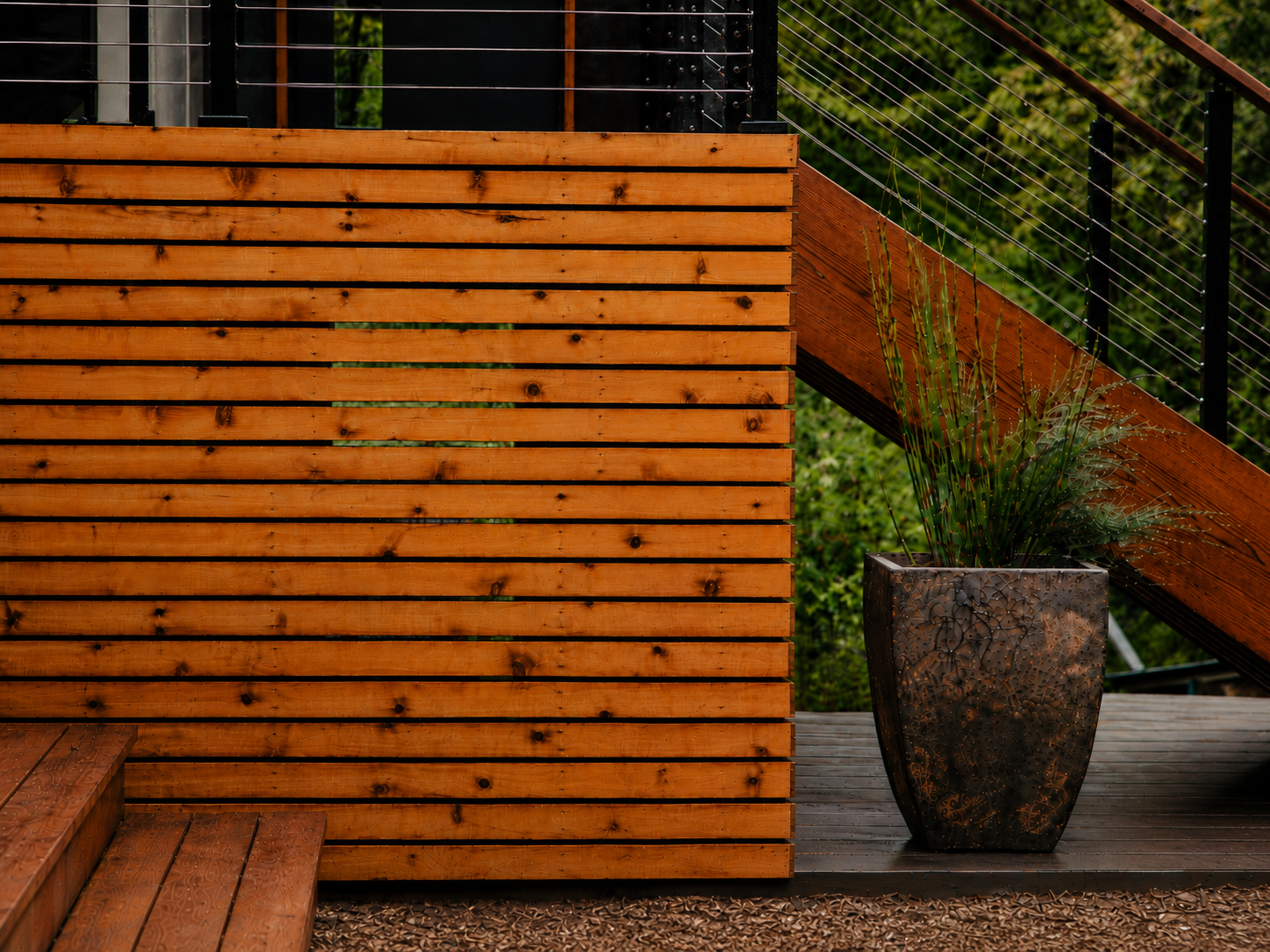 Close-up of a modern outdoor deck with wooden stairs, a wooden privacy wall, a potted plant in a decorative black planter, and a metal cable railing with a diagonal wooden support, surrounded by greenery.