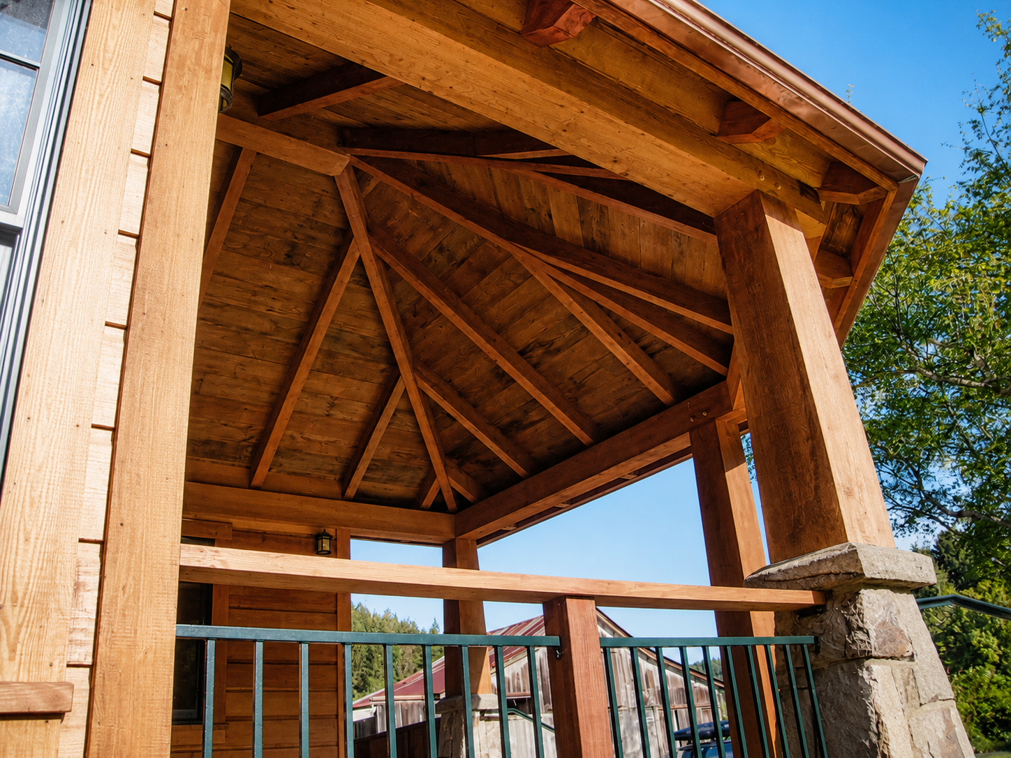 View of a wooden porch or balcony with a stone pillar, supporting a roof with exposed wooden beams, against a backdrop of a clear blue sky and green trees.