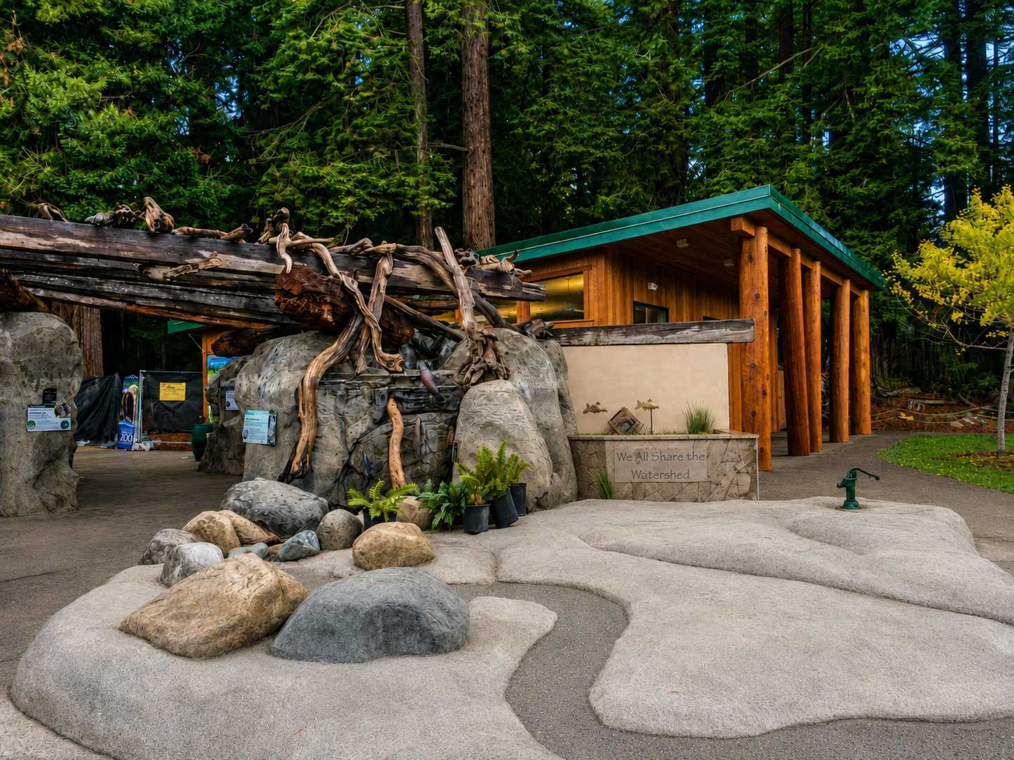 A nature-themed area with rocks, driftwood, plants, a wooden building, and a sign that says "We All Share the Watershed."