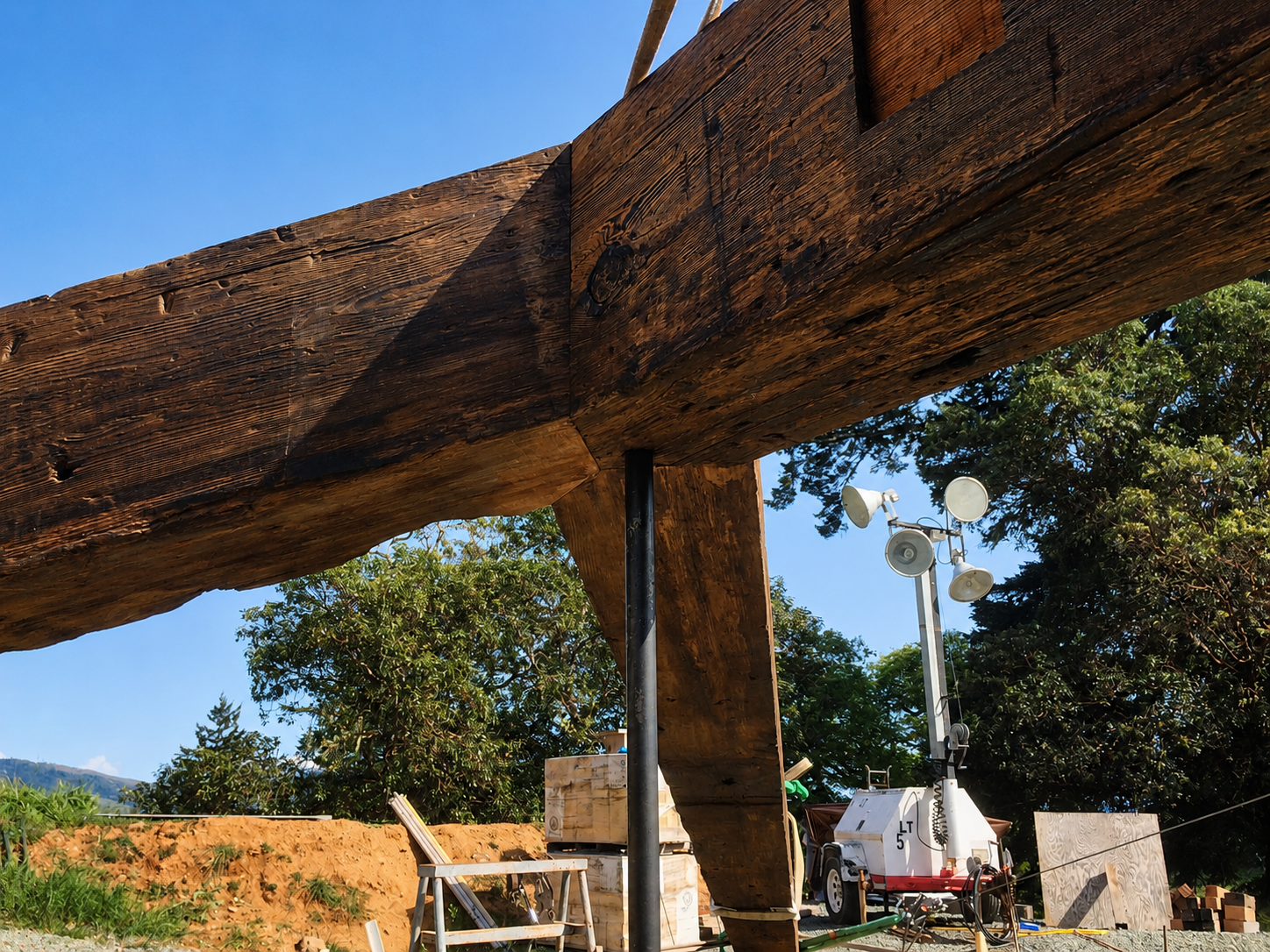 Construction site with large wooden beams, trees in the background, and construction equipment including a mobile light, a boom lift, and other machinery.