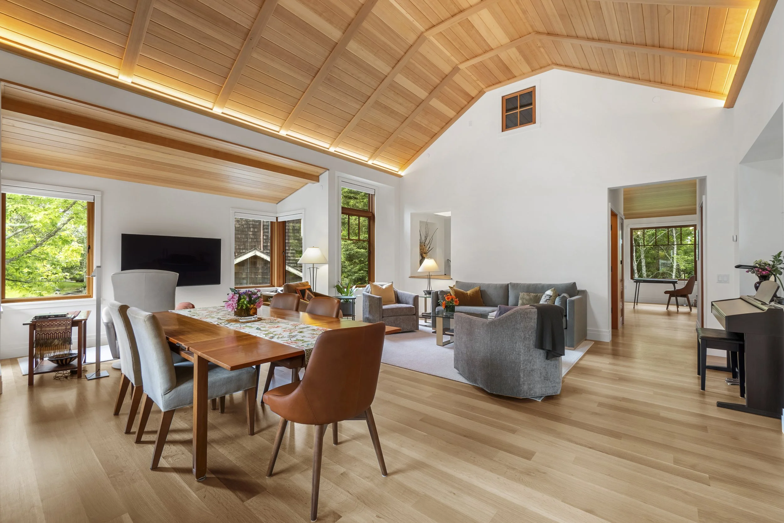Living room with wooden vaulted ceiling, large windows showing greenery outside, a dining table with chairs, a gray sofa with pillows, a TV mounted on the wall, and various lamps and decorative plants.