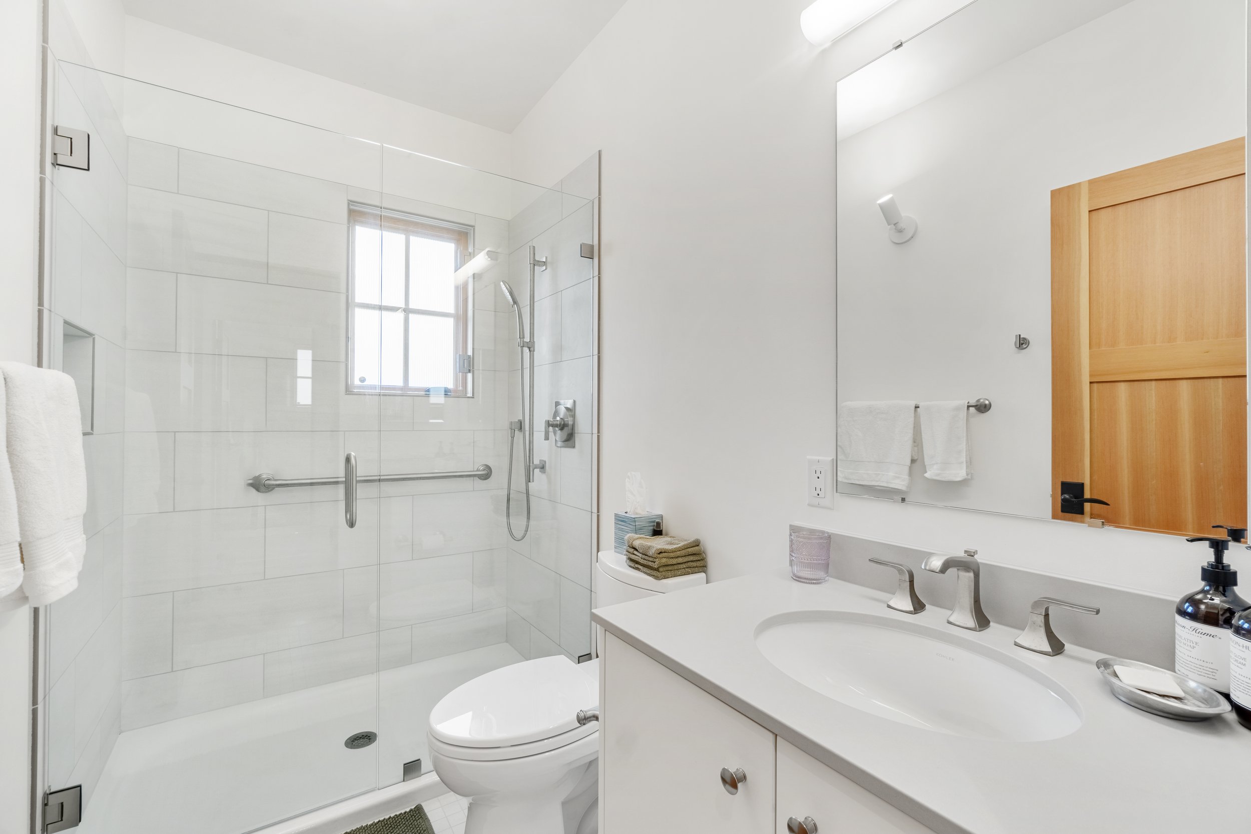 A modern bathroom with a walk-in shower with glass doors, a window, a white vanity with a sink, a mirror, and wooden cabinet doors. Towels and bathroom accessories are visible.