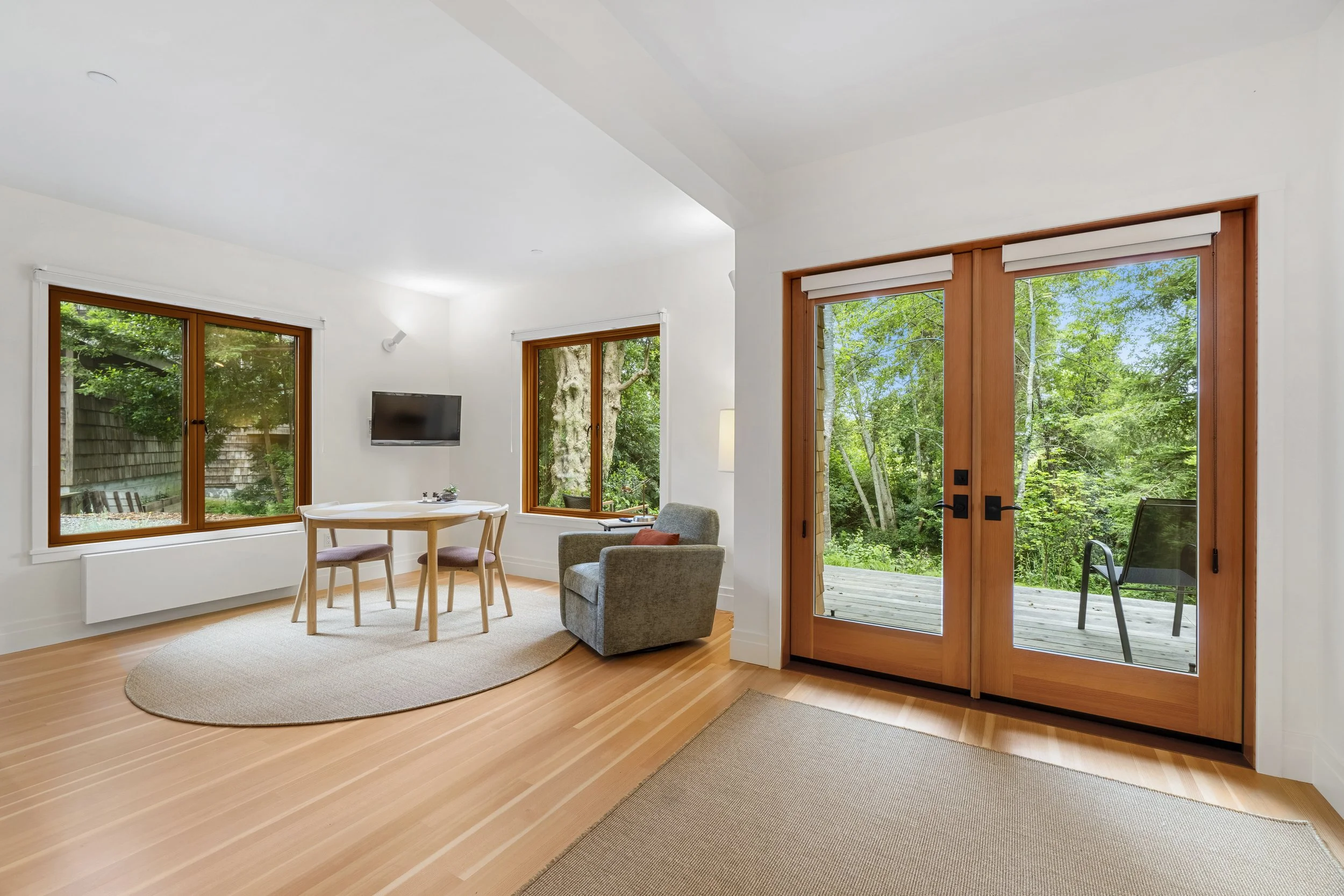 A living room with large windows and sliding glass doors revealing a green outdoor scene, wooden flooring, a round dining table with chairs, a gray armchair, and a wall-mounted flat-screen TV.
