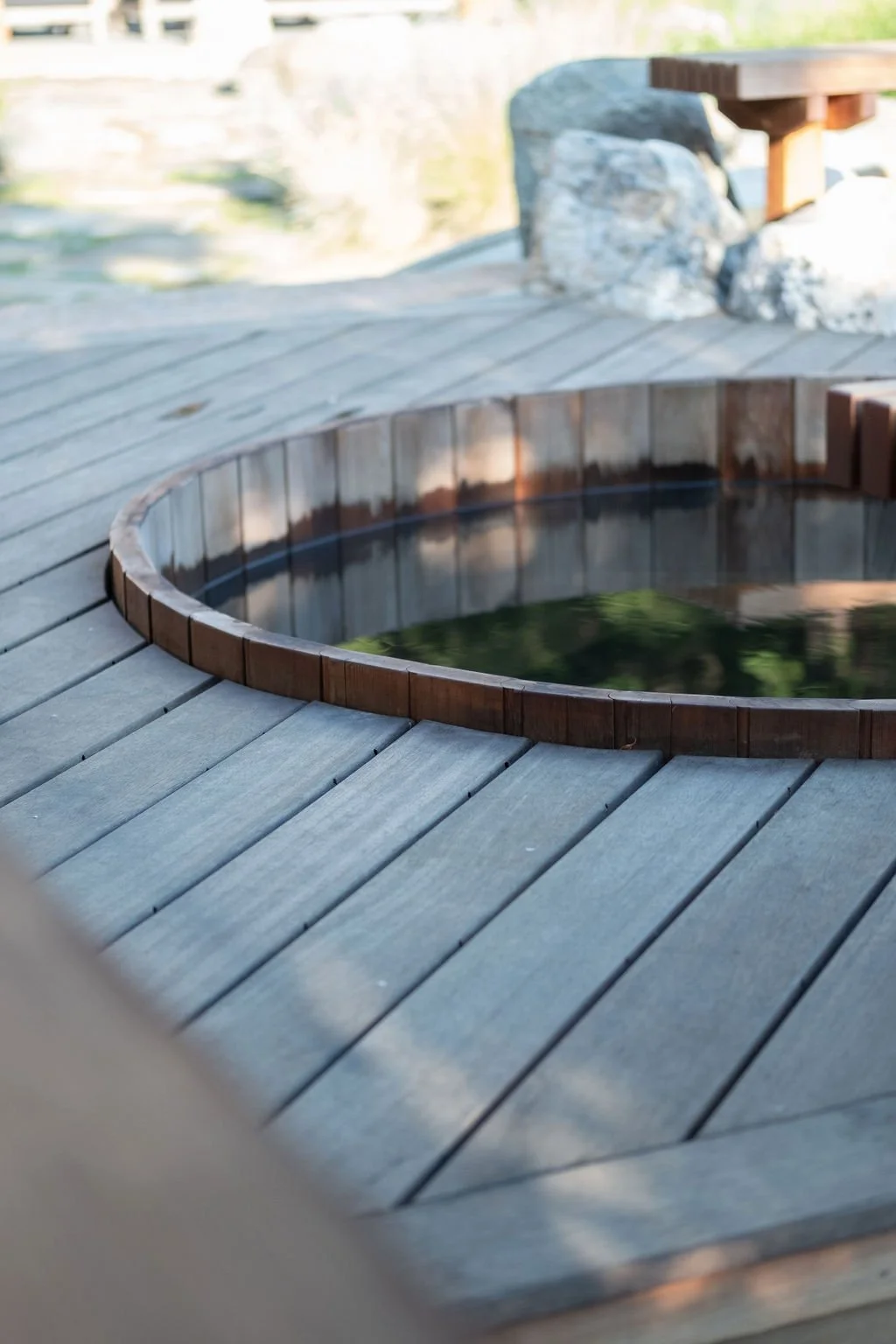 A wooden hot tub with dark water on a wooden deck near a reel with rocks and a bench.