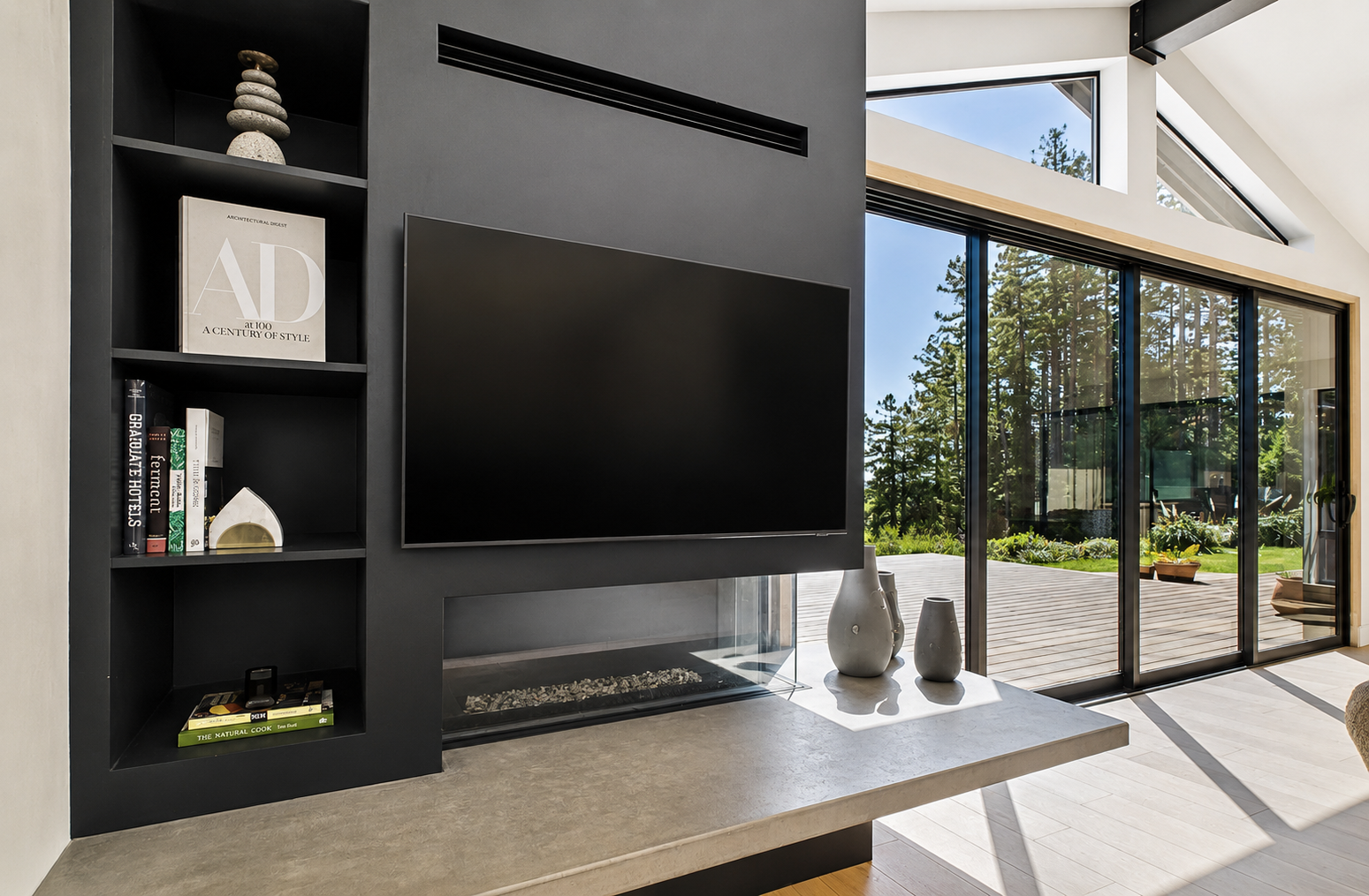 Modern living room with a black wall-mounted TV, a built-in black bookshelf with books and decorative items, a fireplace beneath the TV, and large sliding glass doors leading to an outdoor deck with scenic trees.