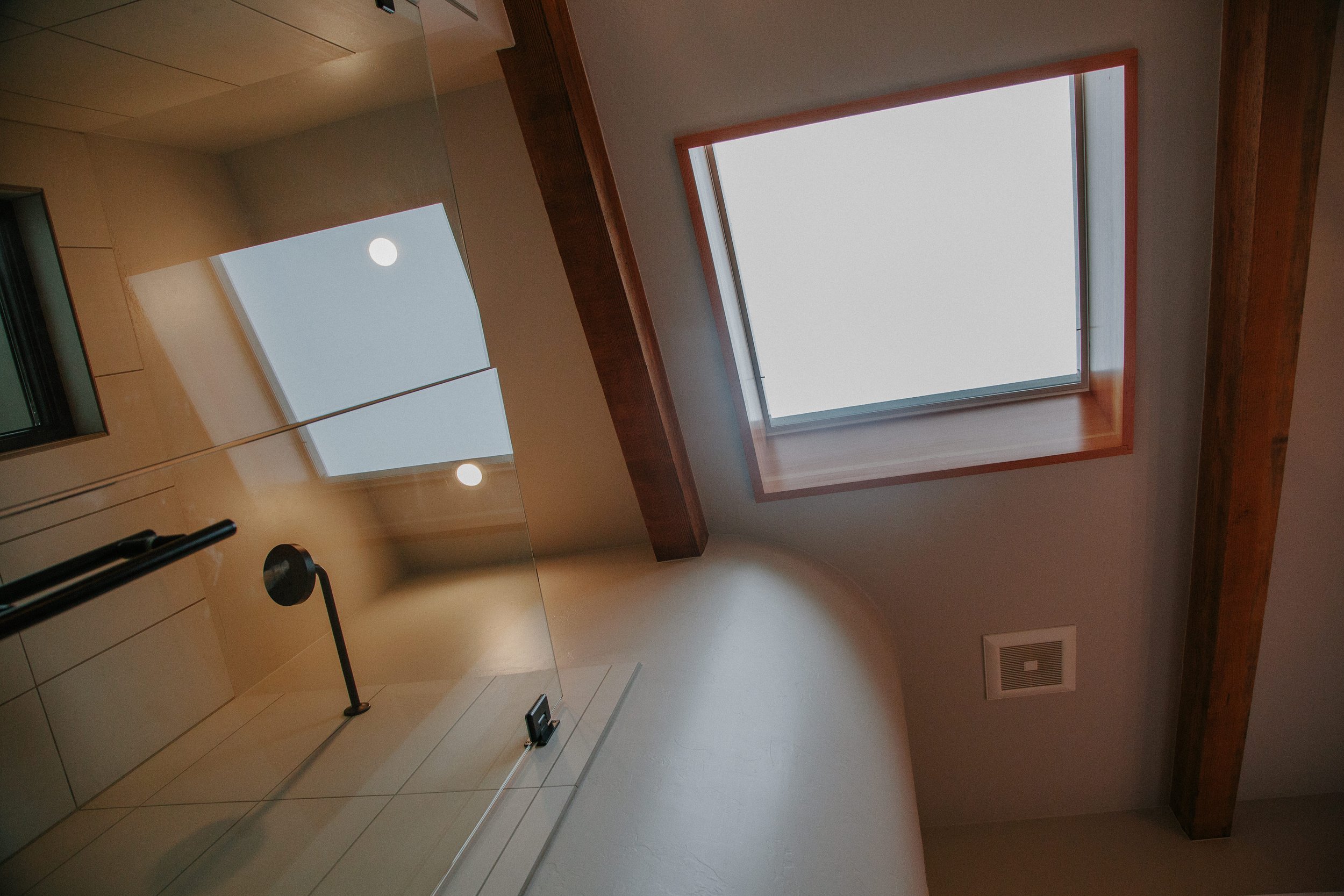 A room with a large skylight window on the ceiling, wooden beams, and a mirror reflecting the skylight. There is a black faucet and tiled wall in the foreground.