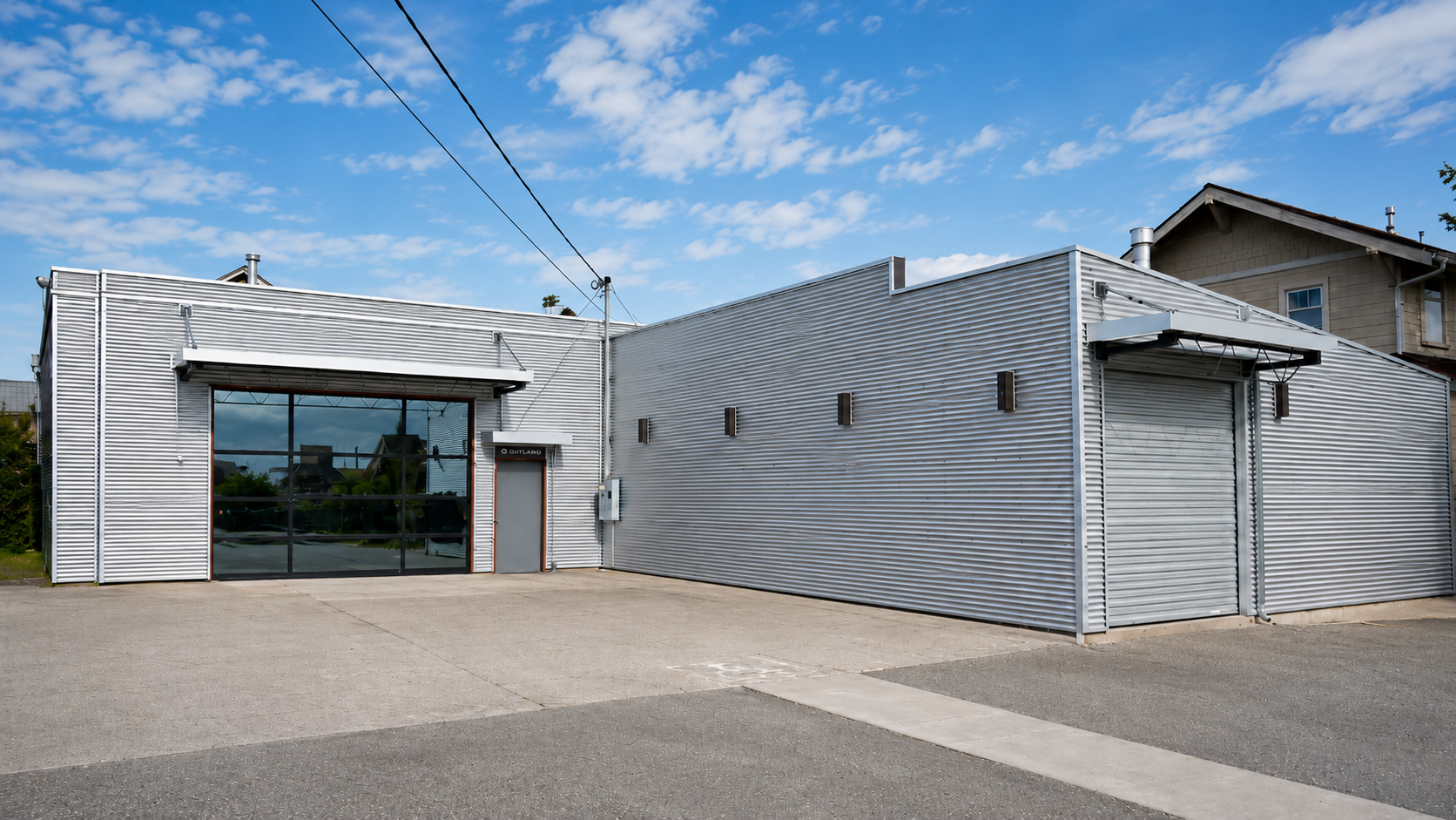 A standalone commercial building with a metal corrugated exterior, large glass garage door, and a smaller door labeled 'OUTLAND,' situated on a paved lot under a blue sky with clouds.