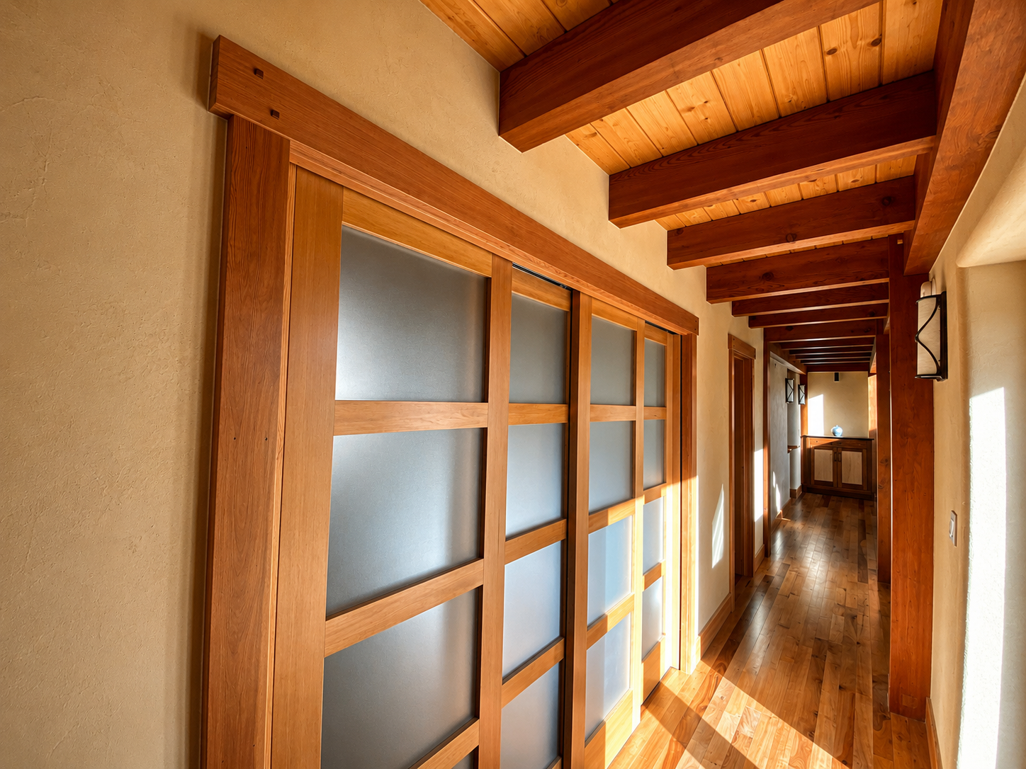 A hallway with wooden beams on the ceiling, a glass door with wooden framing on the left, and sunlight entering through a window on the right. The flooring is hardwood, and there is a small cabinet with a decorative item at the end of the hall.
