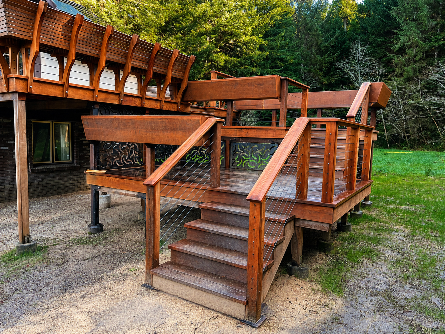 A wooden deck with stairs leading down to a grassy yard, surrounded by tall green trees.