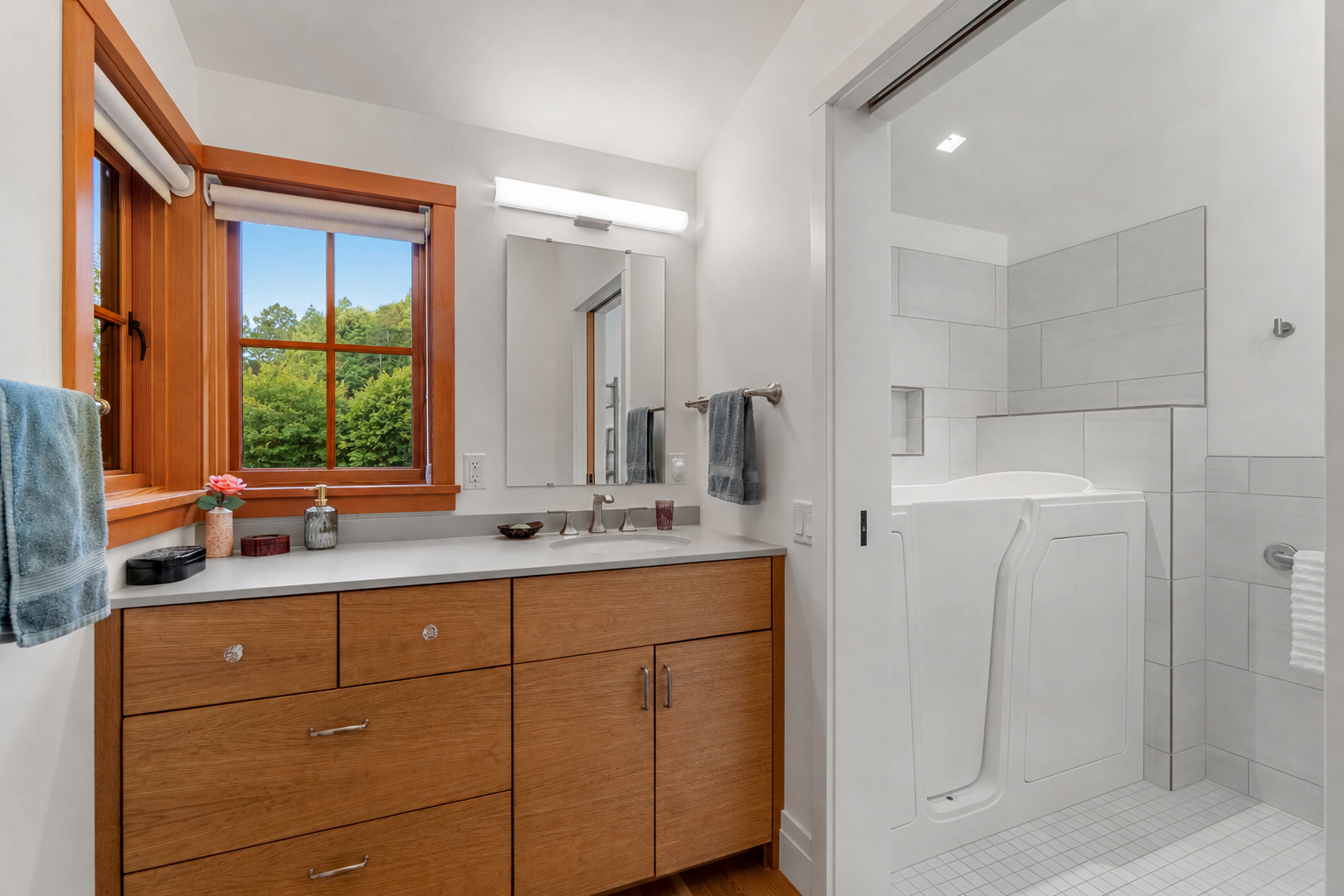 A bathroom with a wooden vanity and a window showing green trees outside. There is a mirror and a shower with a grab bar and tiled walls.