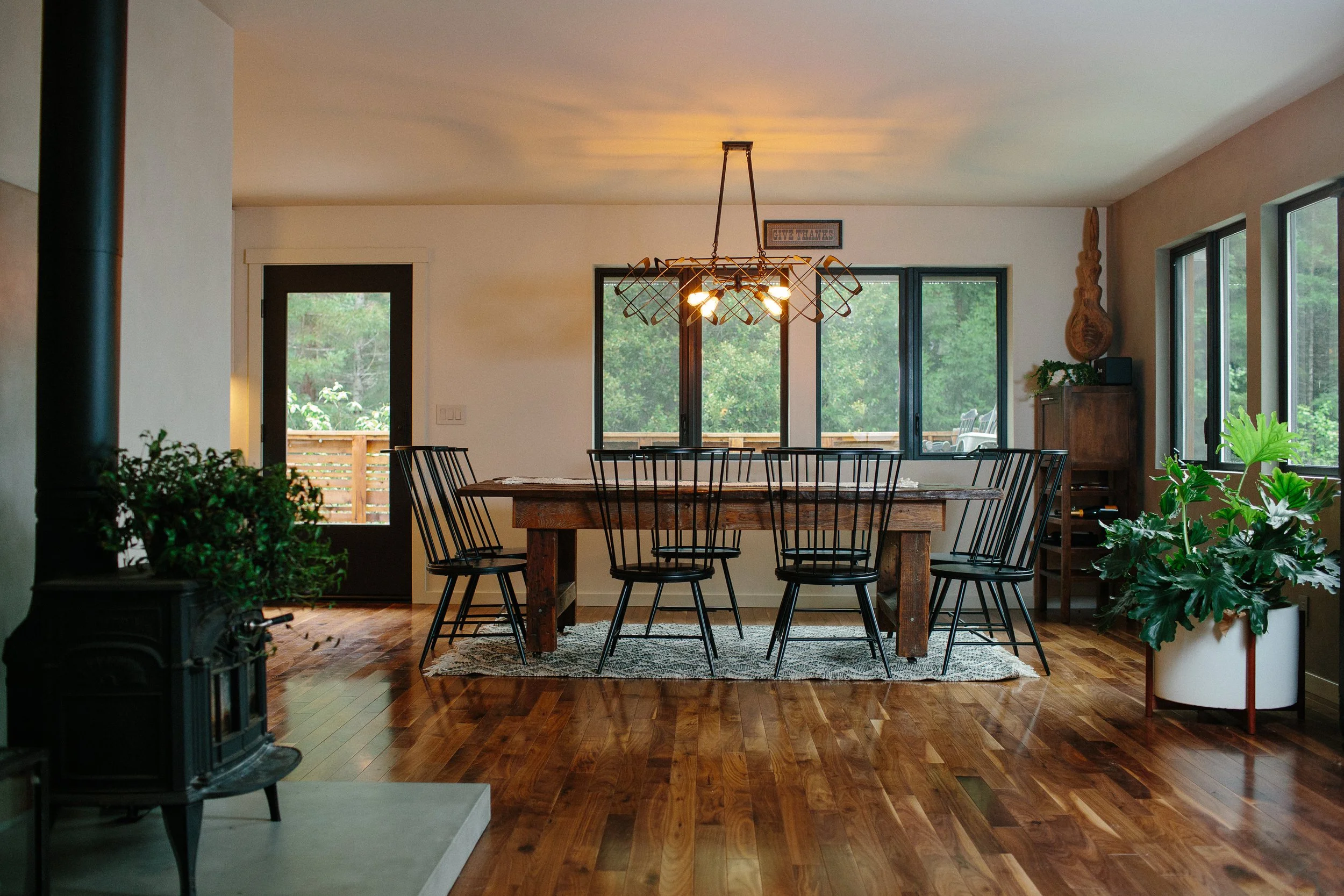 A dining room with a rustic wooden table, black chairs, and large windows showing greenery outside. There is a chandelier hanging above the table, a sign with the words "Give Thanks," and various plants inside the room.