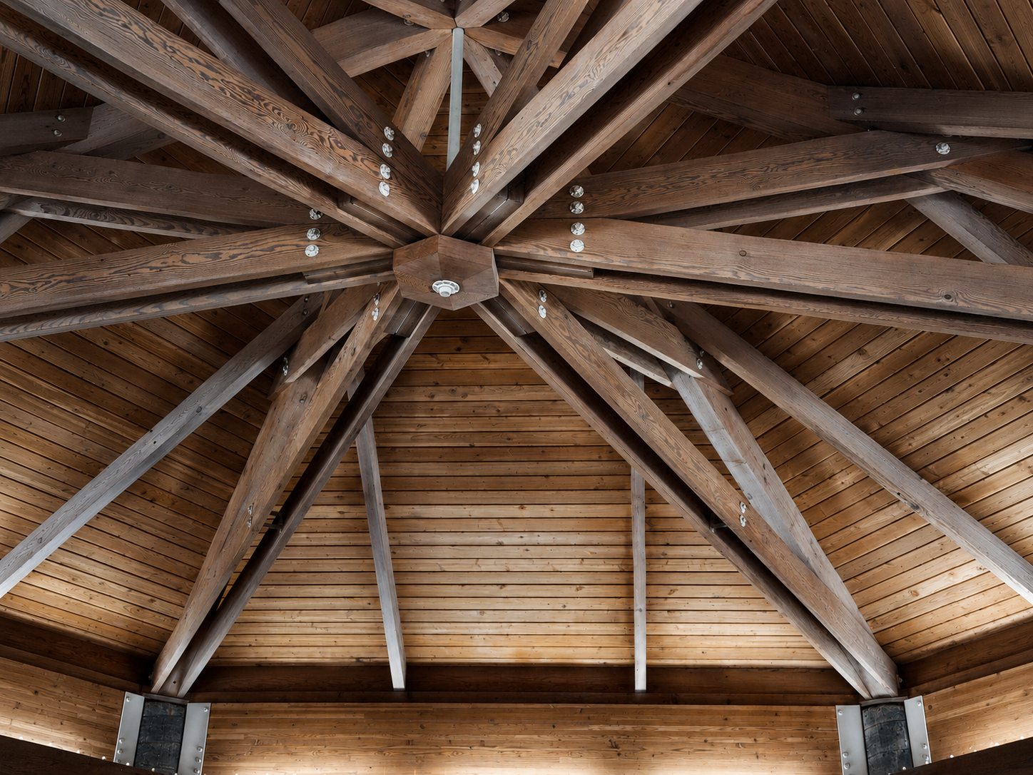 Interior view of a wooden ceiling with large exposed wooden beams arranged in a radial pattern. The ceiling is finished with horizontal wooden planks.