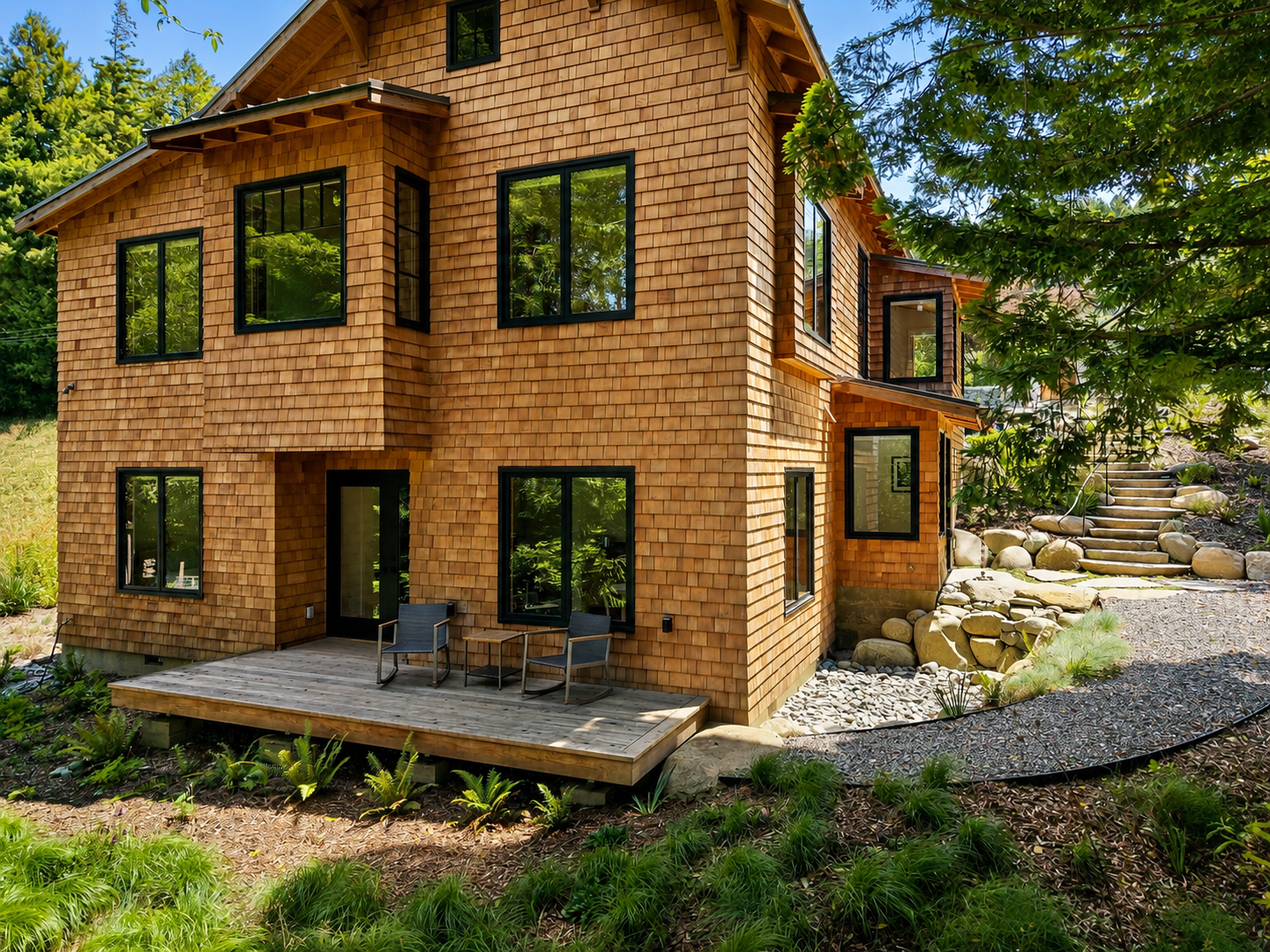 A two-story wooden house with shingle siding and black window frames, set amidst lush greenery and trees. There is a small wooden deck with outdoor chairs in the front.
