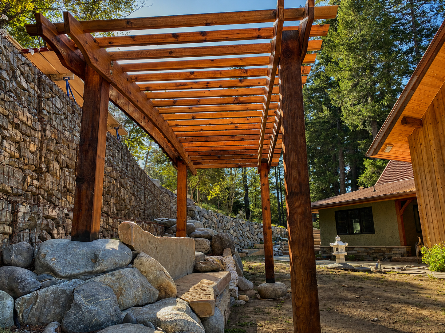 Construction of a wooden pergola in a backyard with rocks and a stone wall on one side, surrounded by trees and a house with a brown roof in the background.