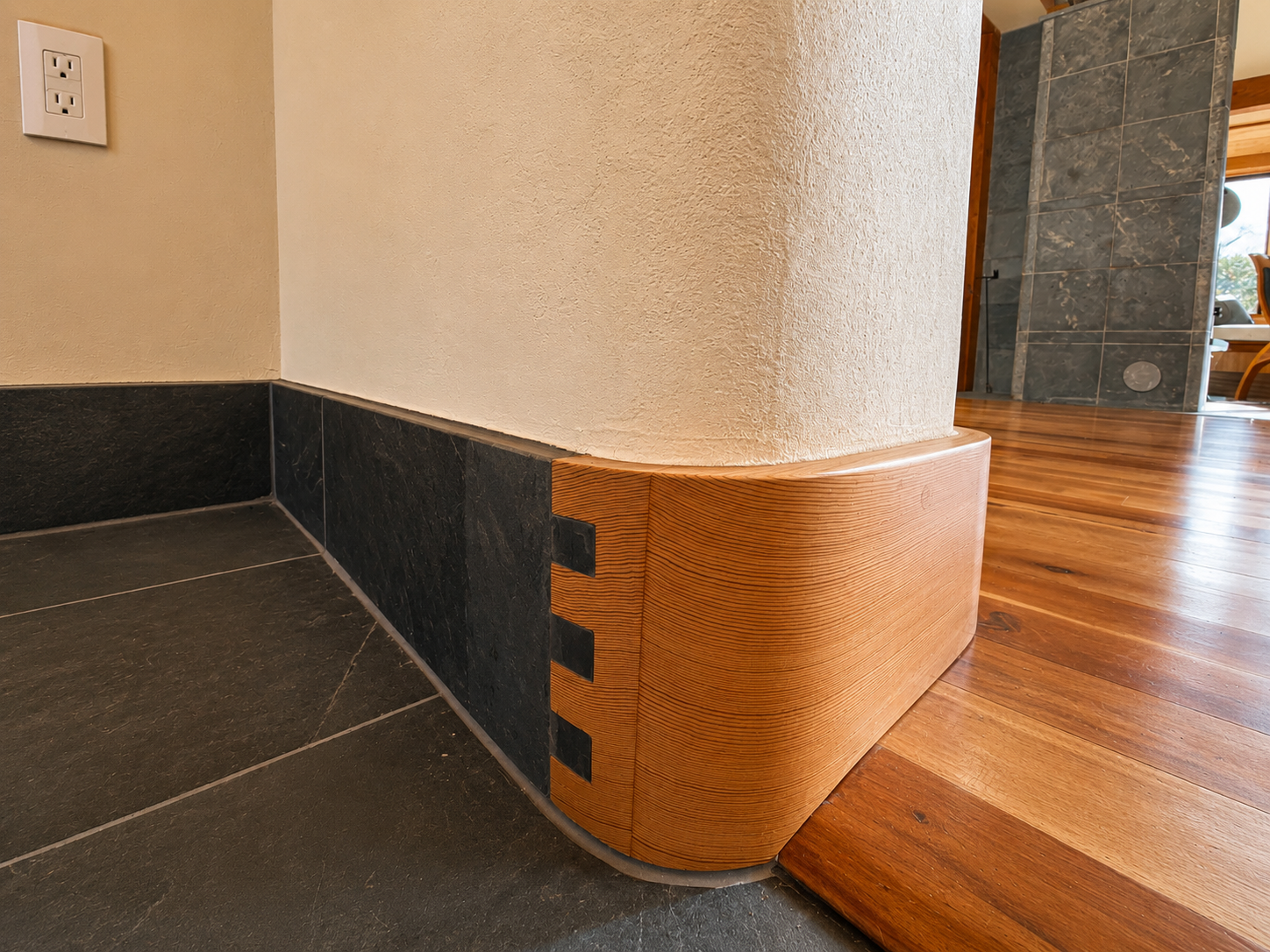 Close-up of a corner of a room showing black tile flooring, a beige textured wall, dark tile baseboard, and a curved wooden molding at the transition between the wall and polished hardwood floor.