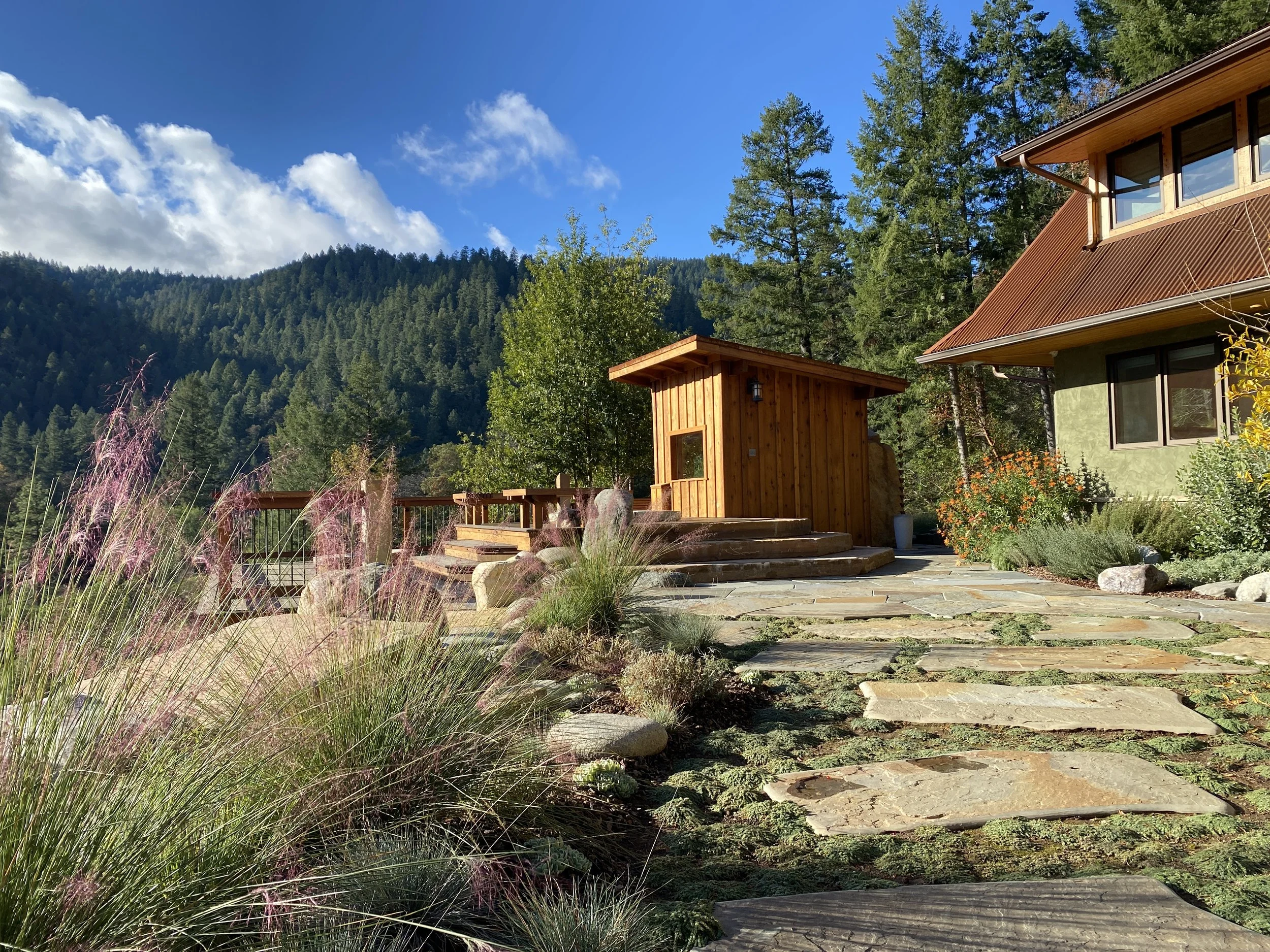 A house with wooden and green exterior walls, set in a mountainous area with evergreen trees. A stone walkway leads to the house, surrounded by ornamental grasses and flowering plants. The sky is mostly clear with some clouds.