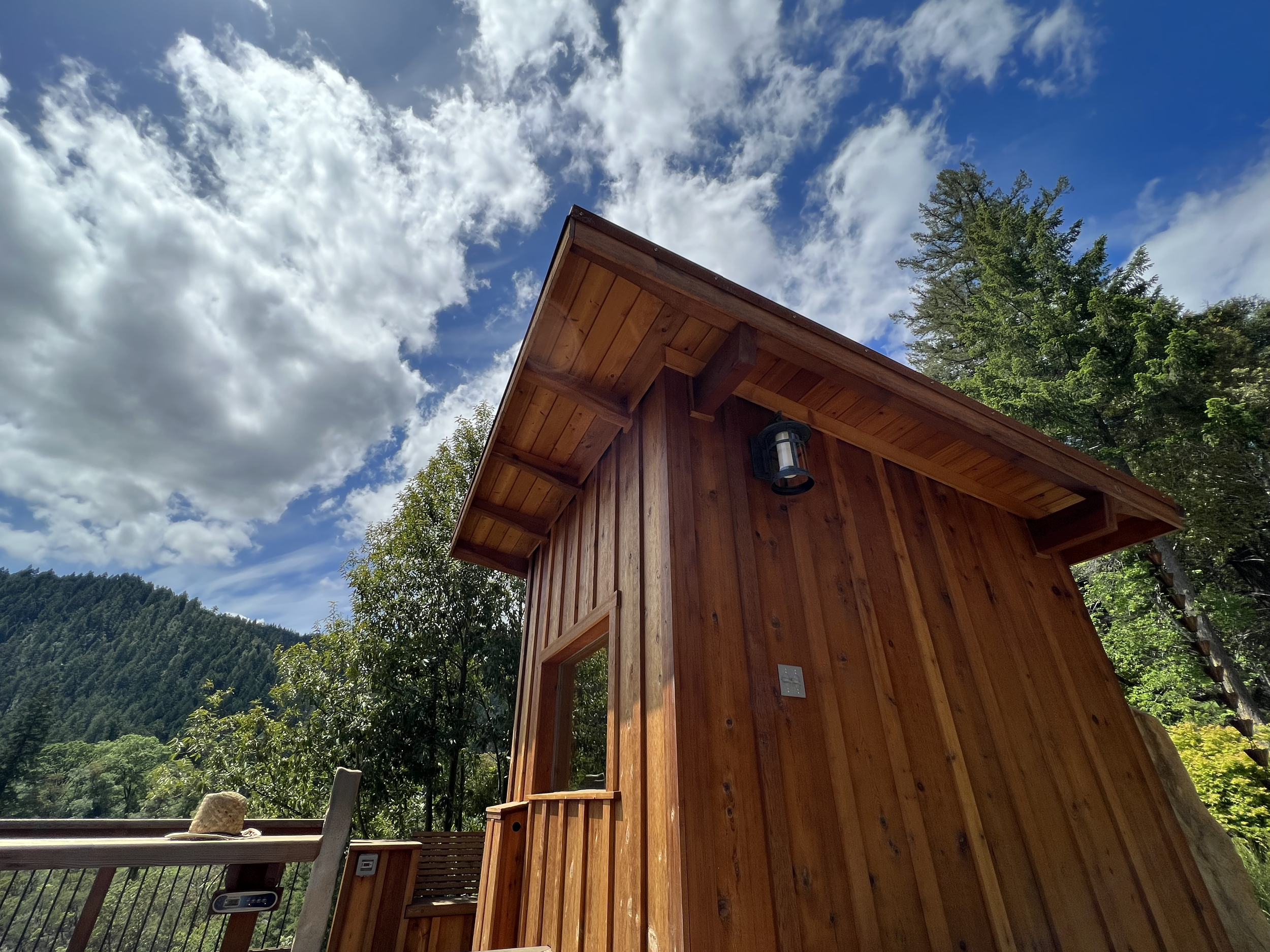 A wooden cabin with an outdoor light fixture against a sky with scattered clouds, surrounded by green trees and mountains.
