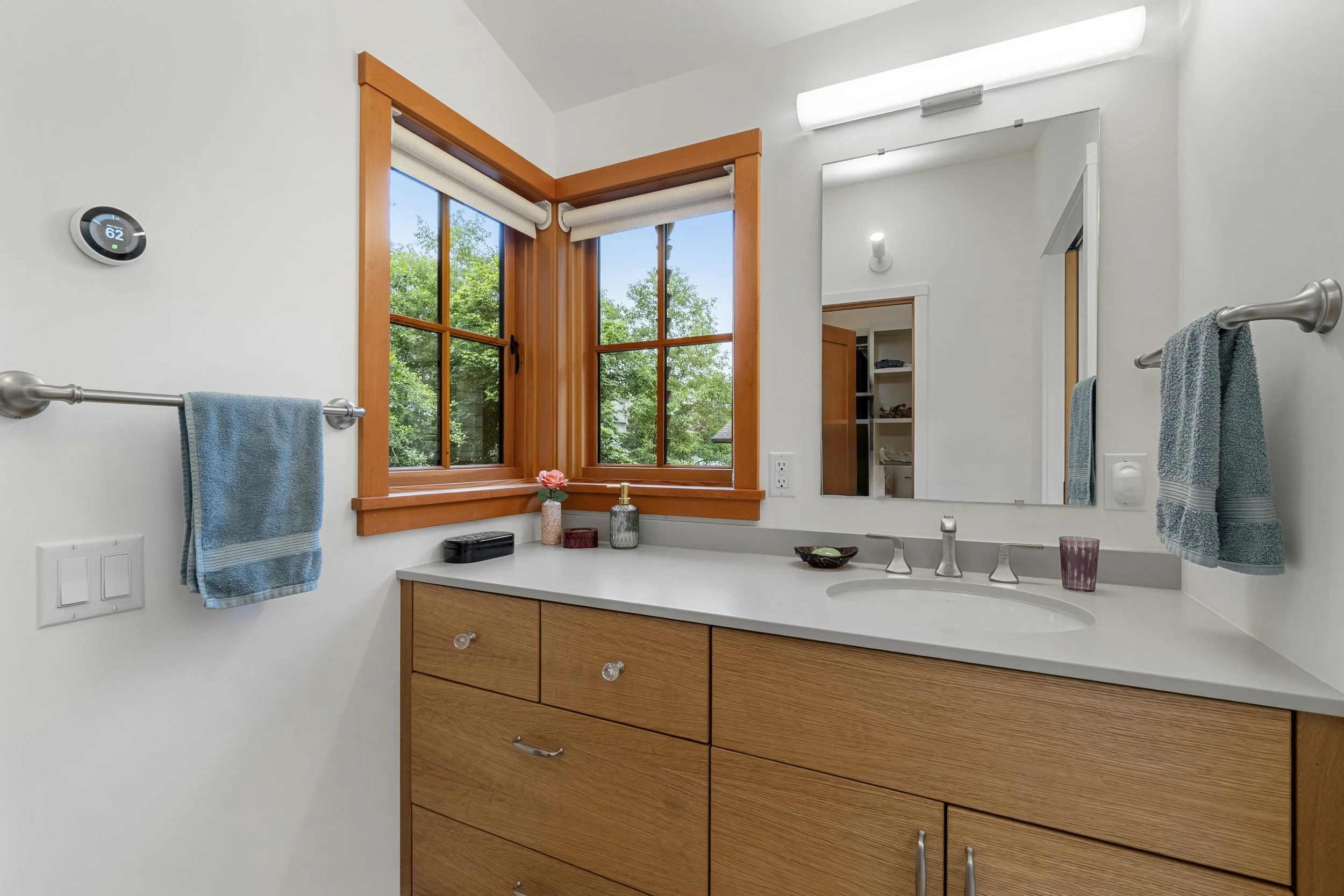 Bathroom vanity with wooden drawers, a white countertop, a small sink, a faucet, and decorative items including a pink flower, glass bottles, and a bowl, with a mirror, towel bars with blue towels, a window with wooden trim, and a view of green trees