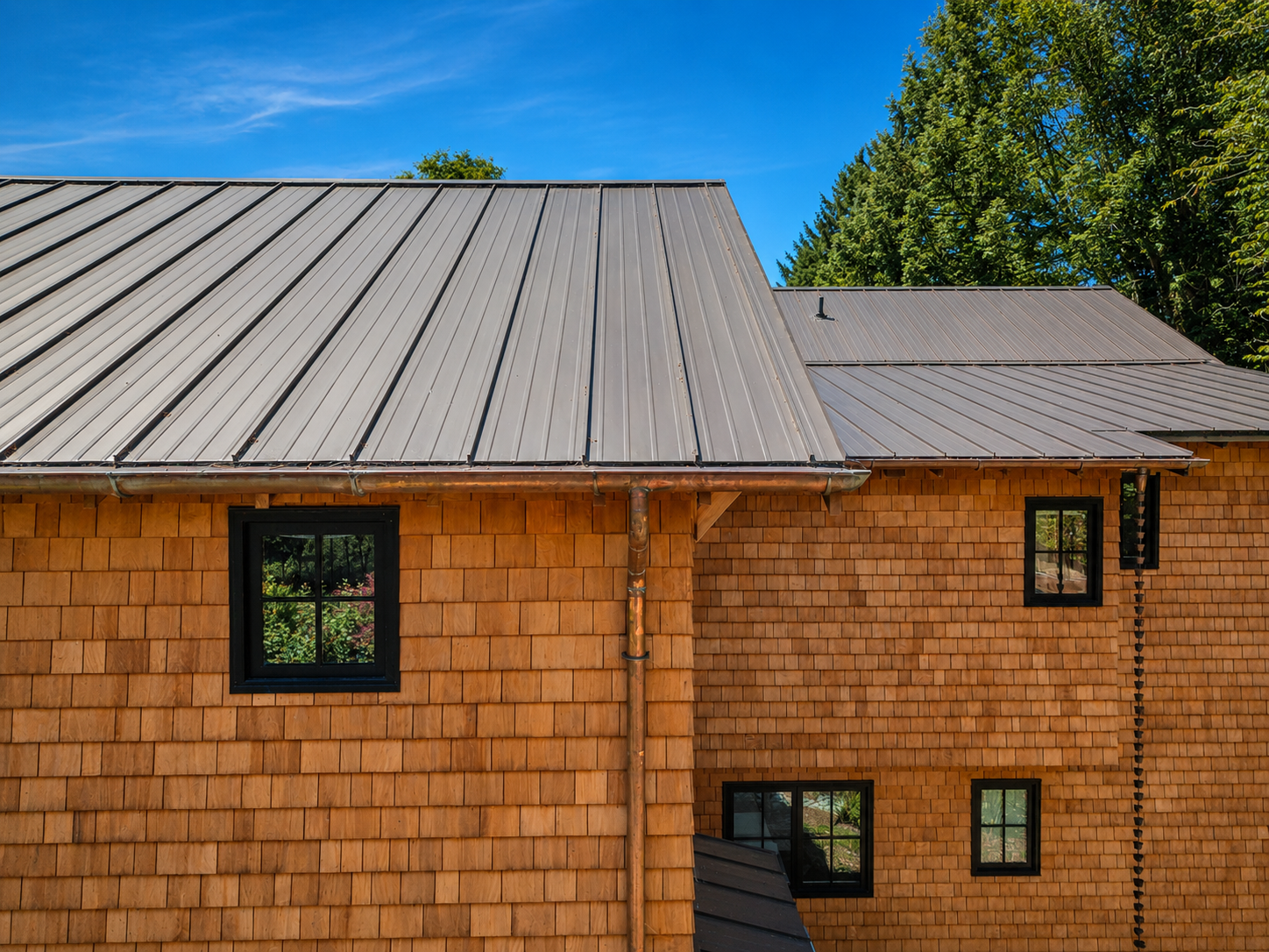 Close-up of a house with wooden clapboard siding, black-framed windows, and a metal roof under a blue sky with trees in the background.