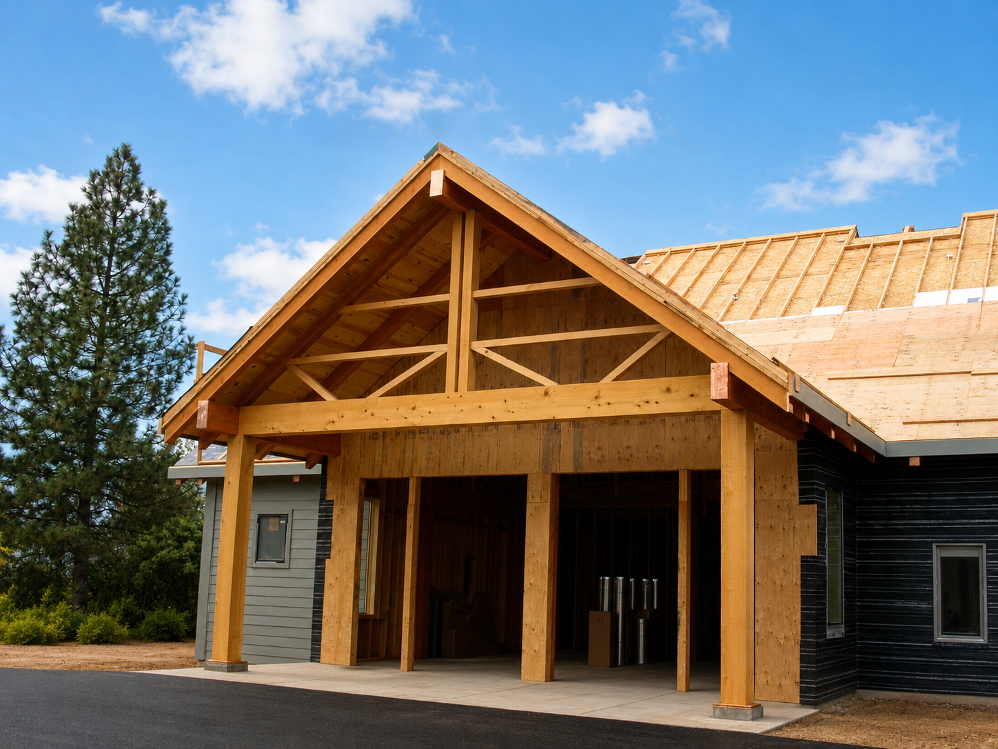 Wooden house under construction with a partially completed roof, green tree, blue sky, and scattered clouds in the background.