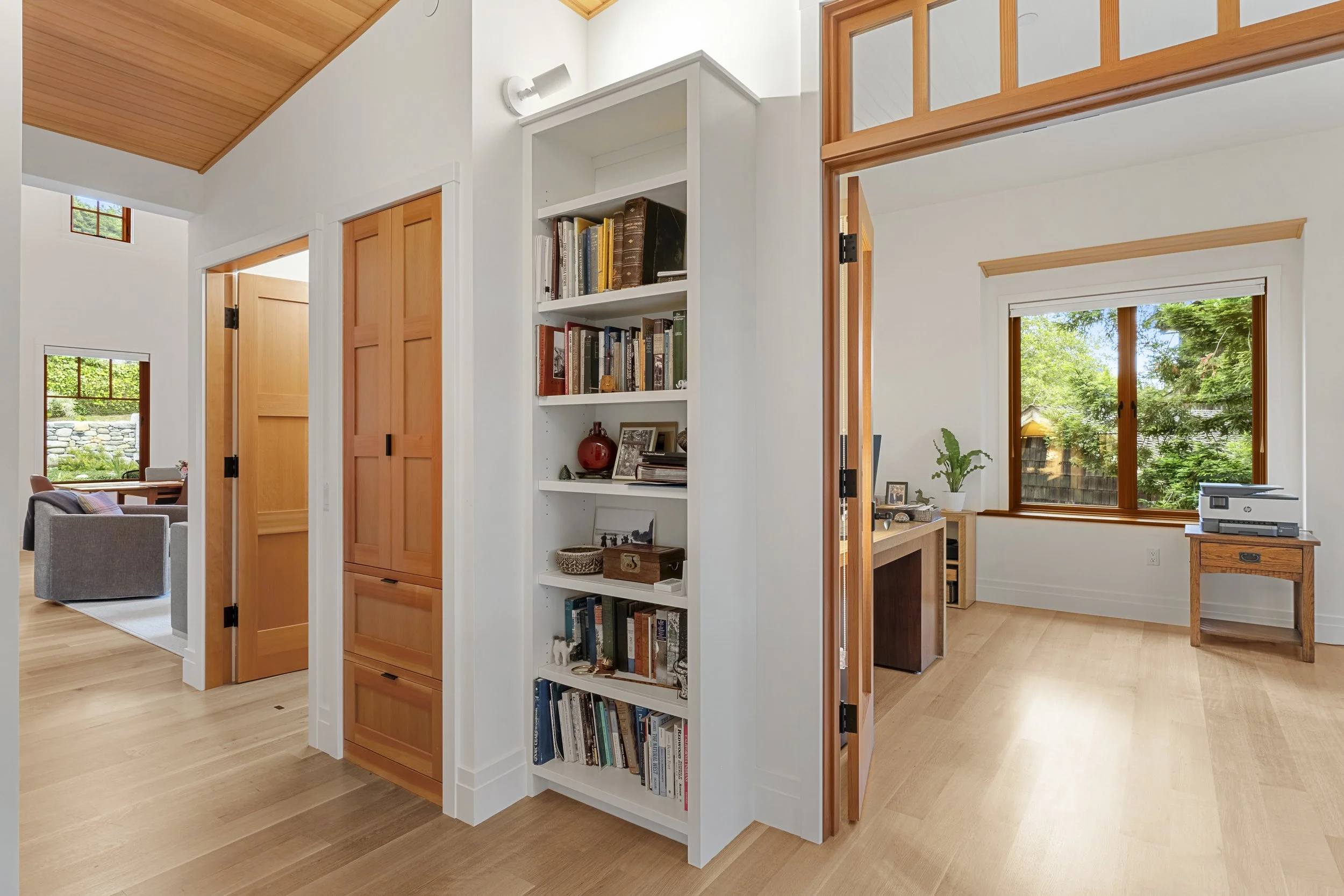 A view of a bright living space with wooden floors and trim, featuring white walls, a built-in bookshelf filled with books and decorative items, and large windows showing green trees outside. There's a small desk with a printer near one window.
