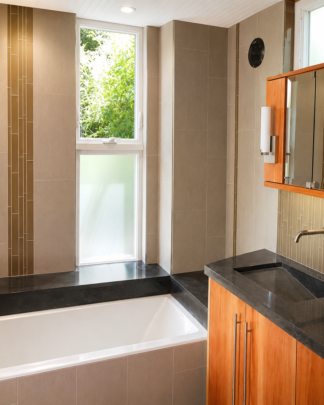 A modern bathroom featuring a bathtub beneath a large window with frosted lower pane and clear upper pane showing greenery outside. The room has beige tiled walls with a decorative vertical strip, a black countertop with a rectangular sink, a wooden 