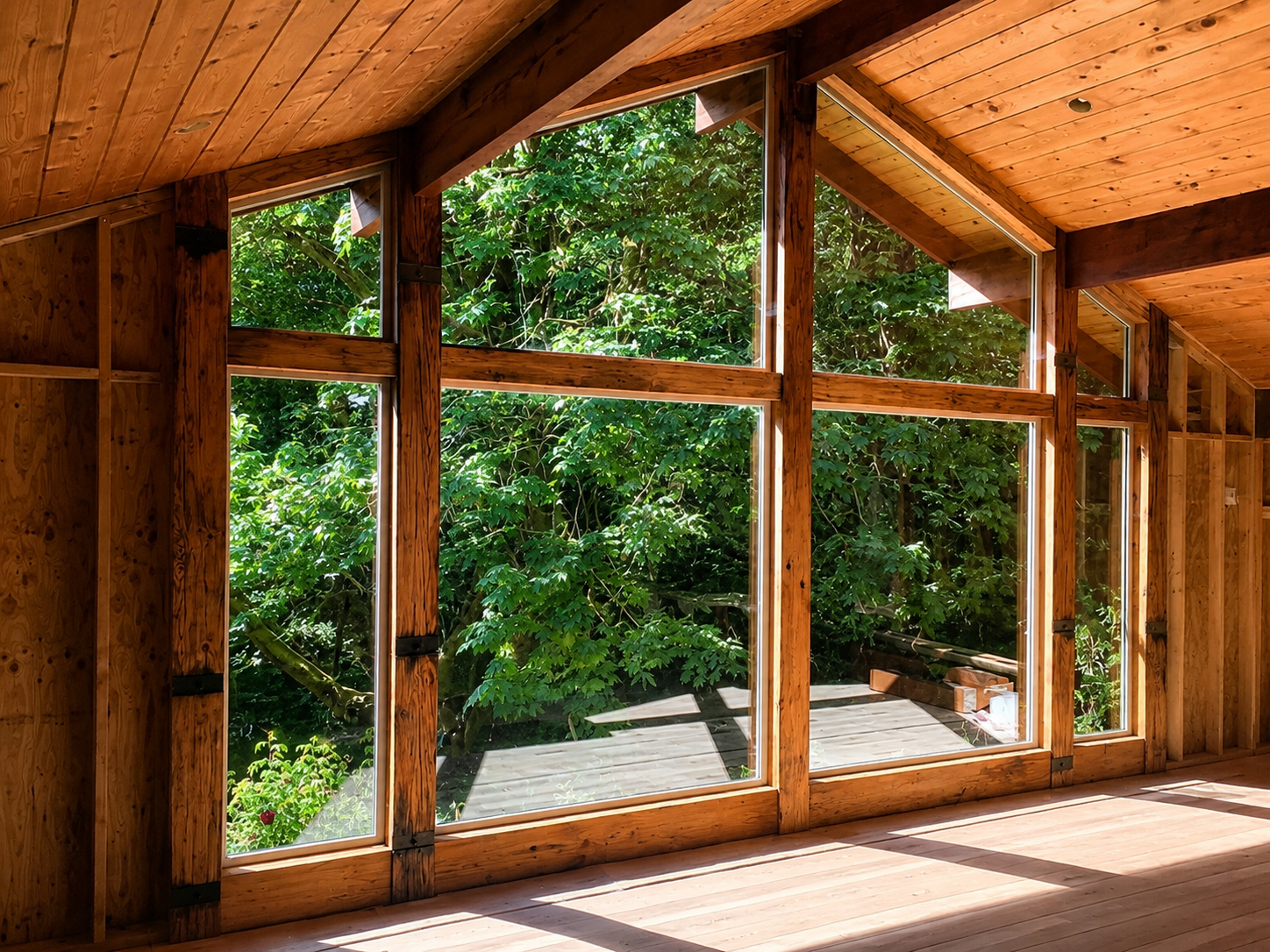 Interior view of a wooden room with large windows that reveal green trees outside, sunlight casting shadows on the wooden floor.