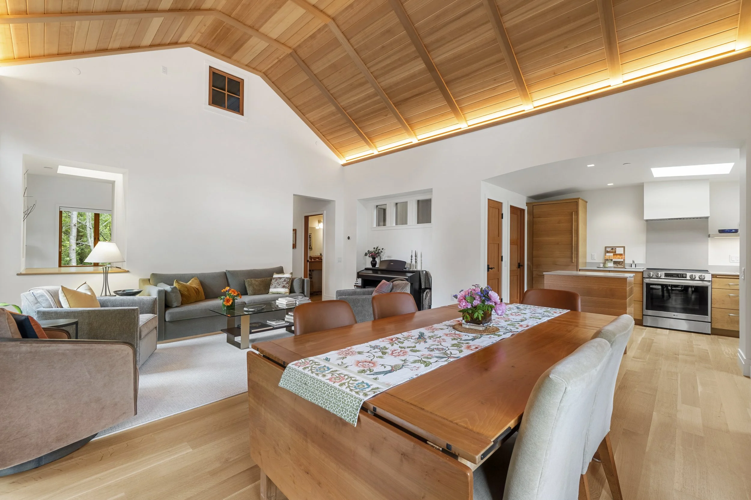 An open-concept living and dining room with a wooden vaulted ceiling, white walls, and hardwood flooring. The living area has sofas, a coffee table, and windows showing greenery outside. The dining area has a wooden table with a floral tablecloth and chairs. The kitchen in the background has wooden cabinets and stainless steel appliances.