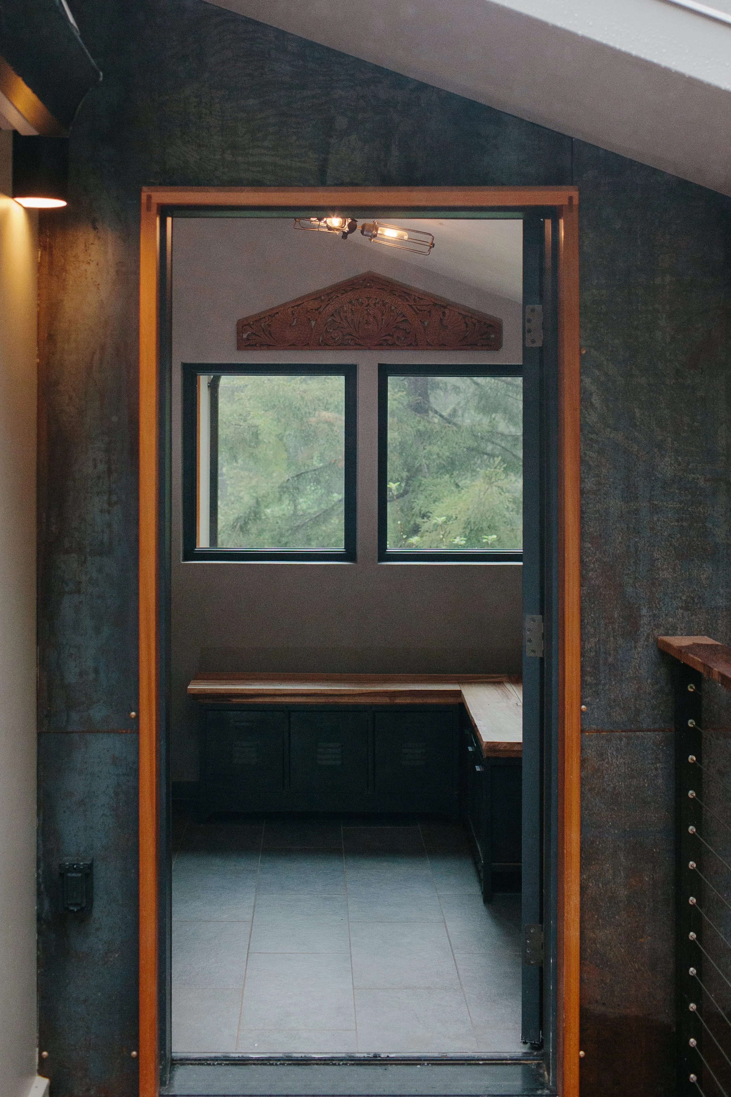 Interior view through a doorway showing a room with a large window, a wooden decorative panel above the window, a black cabinet with drawers, and a tile floor.