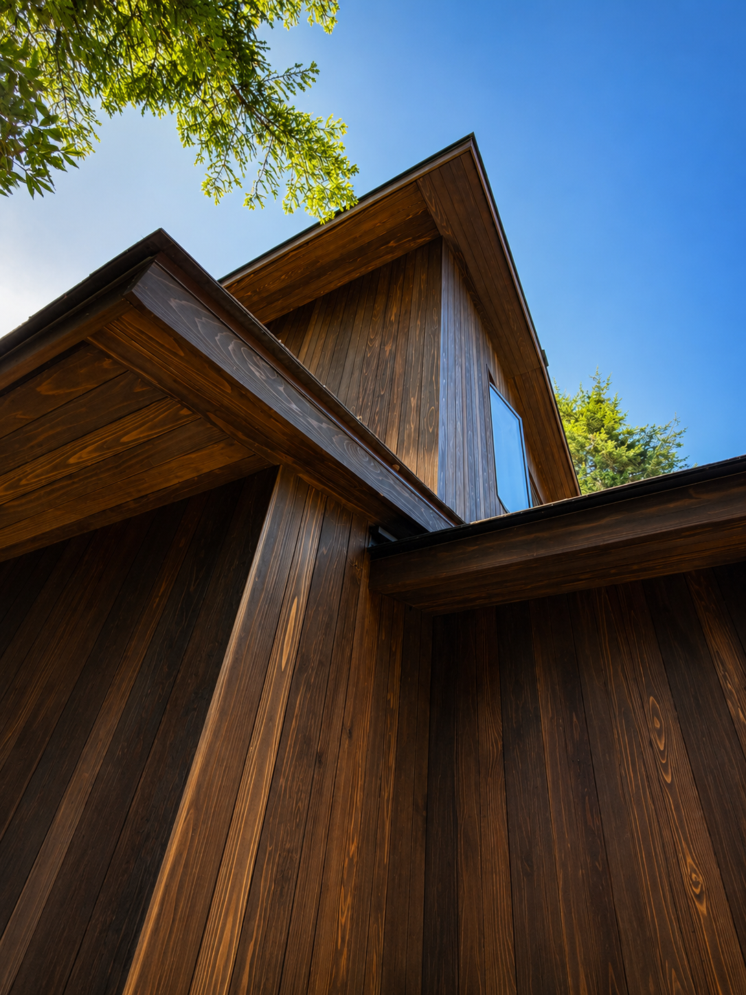 Low-angle view of a modern wooden house with sharp rooflines, blue sky, and green trees in the background.