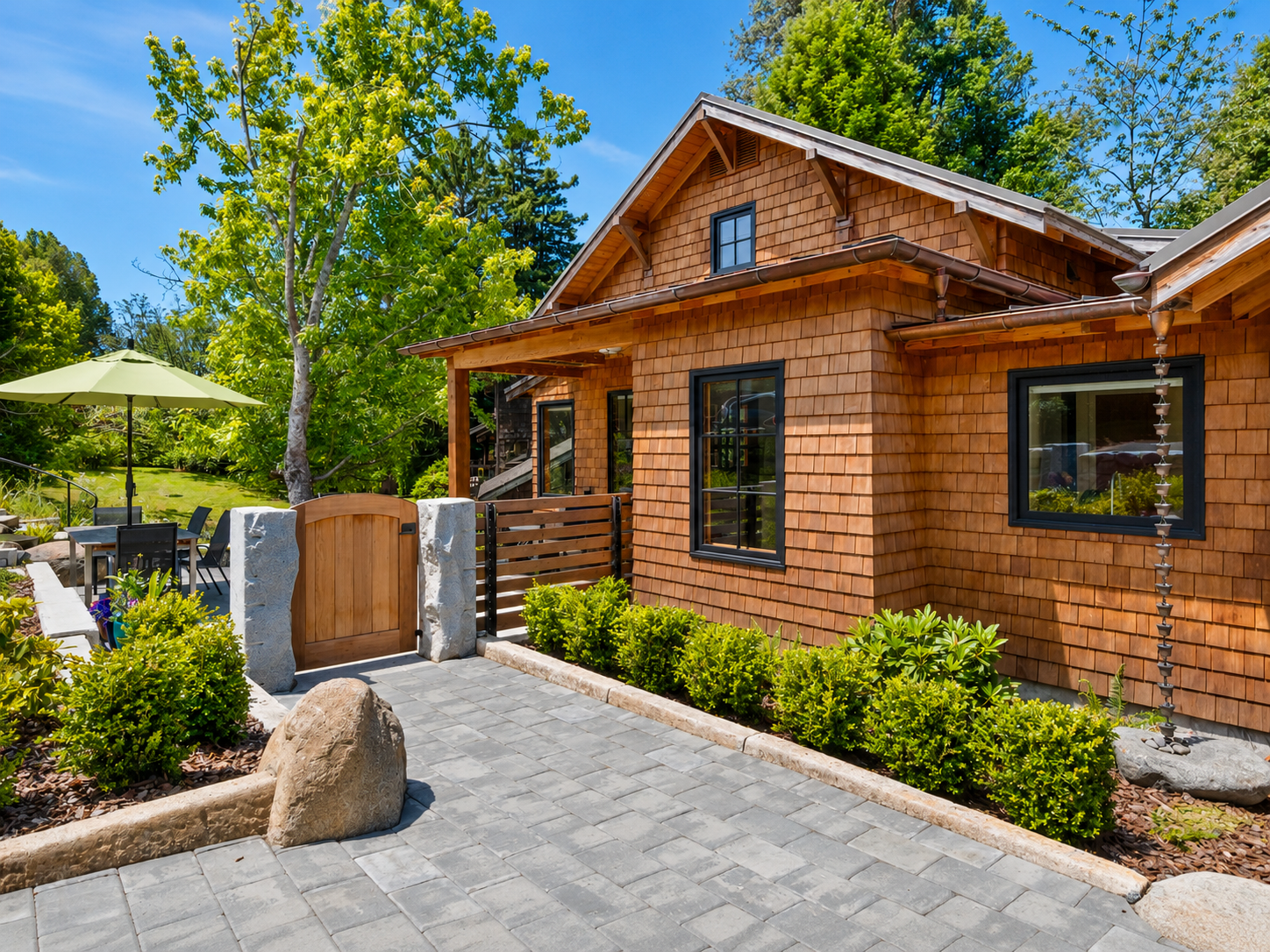 A house with a wood-shingle exterior, black framed windows, and a small front yard with plants, a stone pathway, and a patio area with outdoor furniture and a large green umbrella.