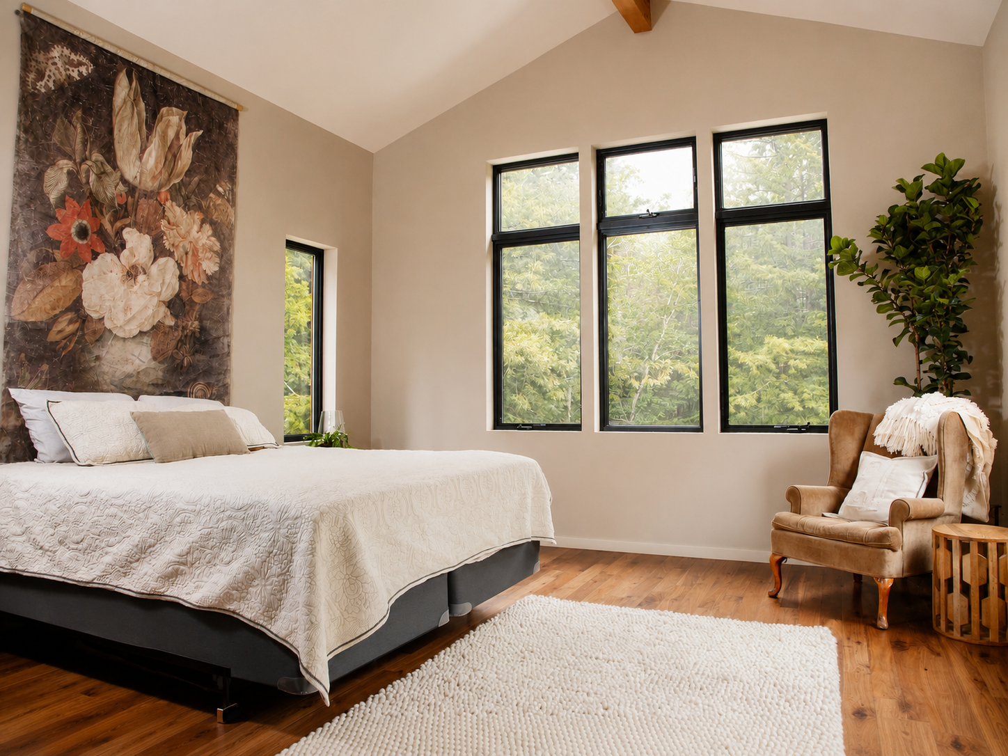 A bedroom with a large bed with white bedding and pillows, a floral tapestry on the wall, three large windows showing green trees outside, a beige armchair with a white pillow and throw, a small side table, a potted plant, and a white textured rug.