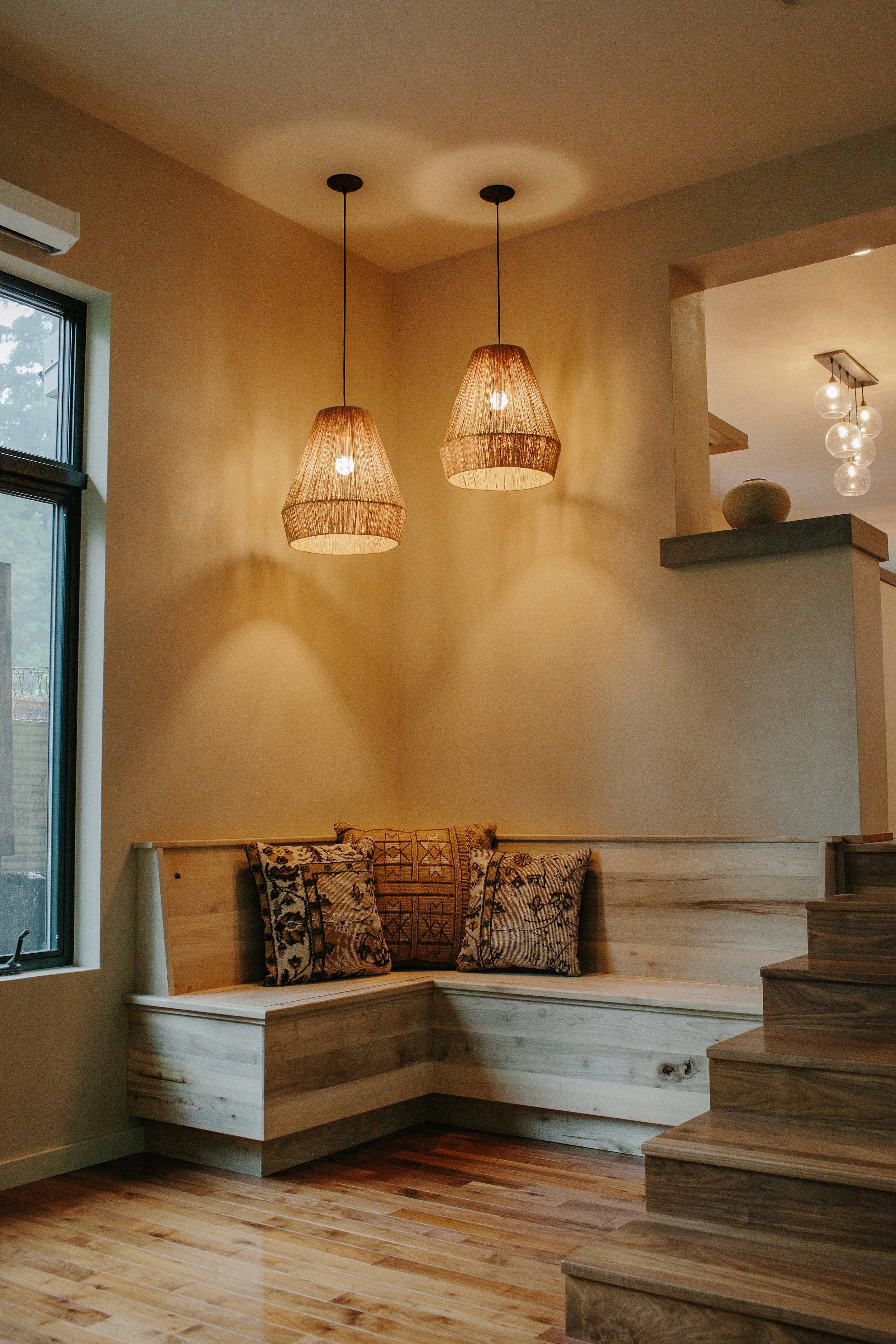 Interior of a room with natural wooden stairs and a built-in corner seating area with patterned cushions. Two pendant lights hang from the ceiling, and there is a window on the left side. A decorative vase is visible on a ledge in a different section