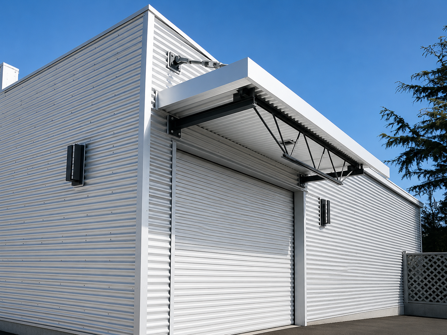 Modern building with metal siding and a closed roller door, featuring an retractable awning and outdoor lighting, under a clear blue sky with a tree visible on the right.